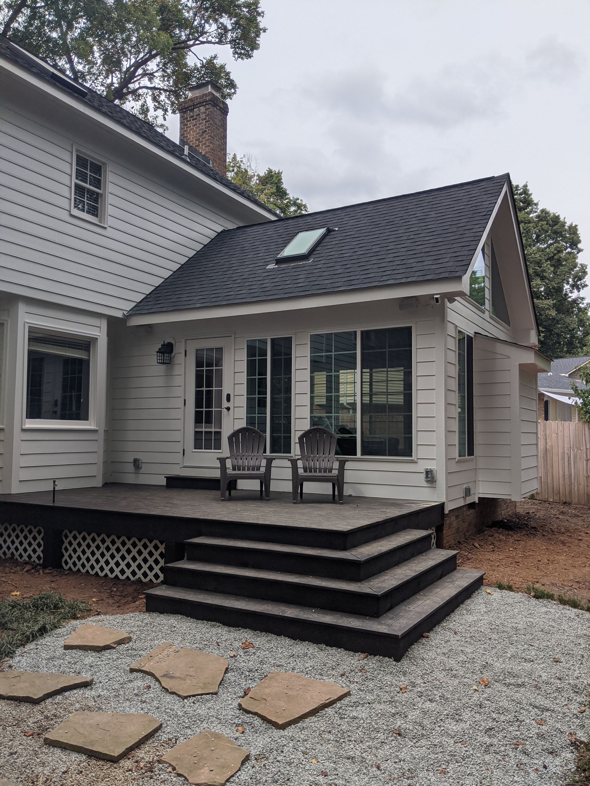 A white two-story house with a rear addition, featuring a dark deck with steps leading to a gravel yard with stone pavers.