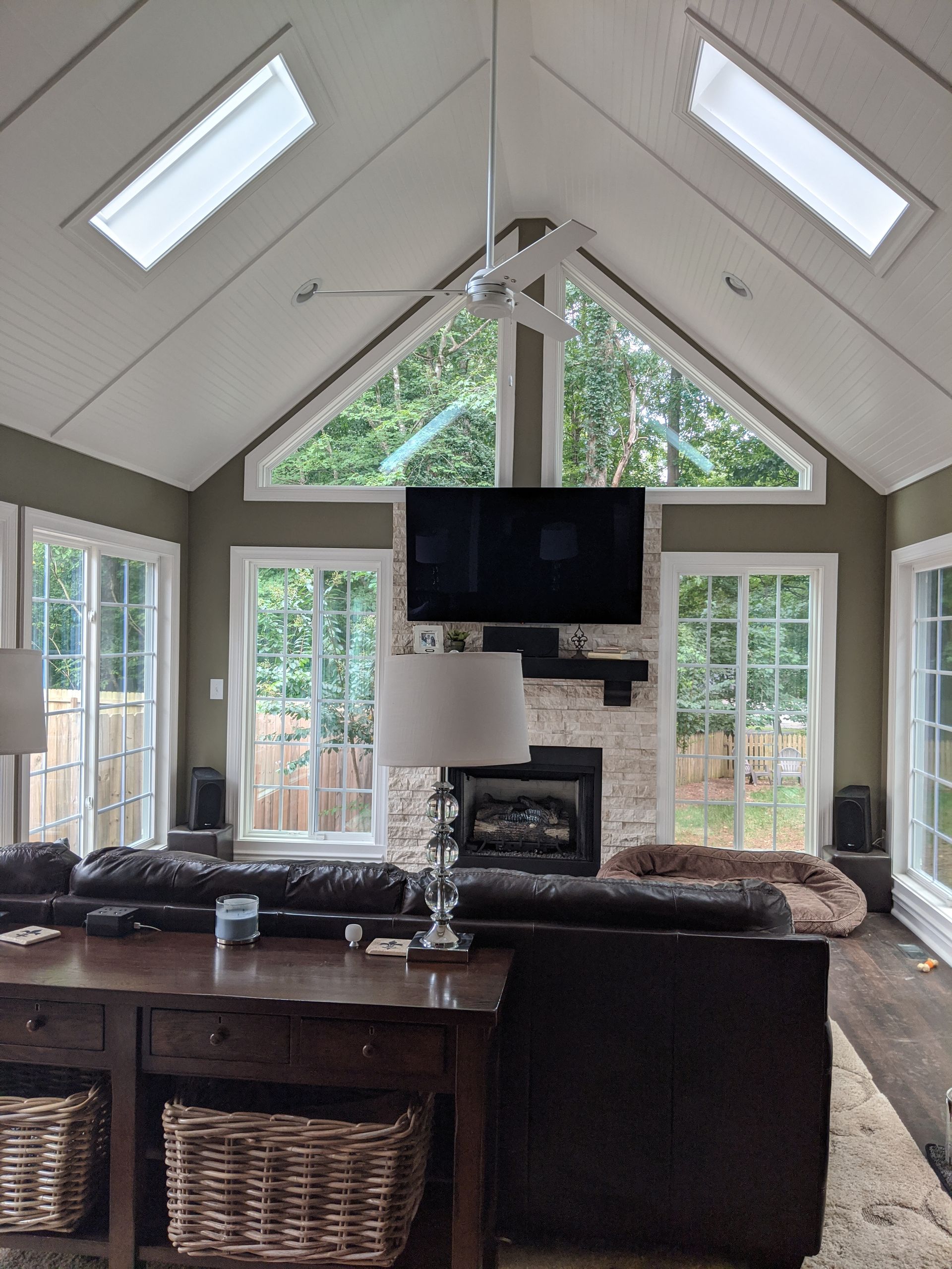 A cozy living room with a vaulted ceiling, skylights, a brick fireplace, a mounted TV, and a leather sofa.