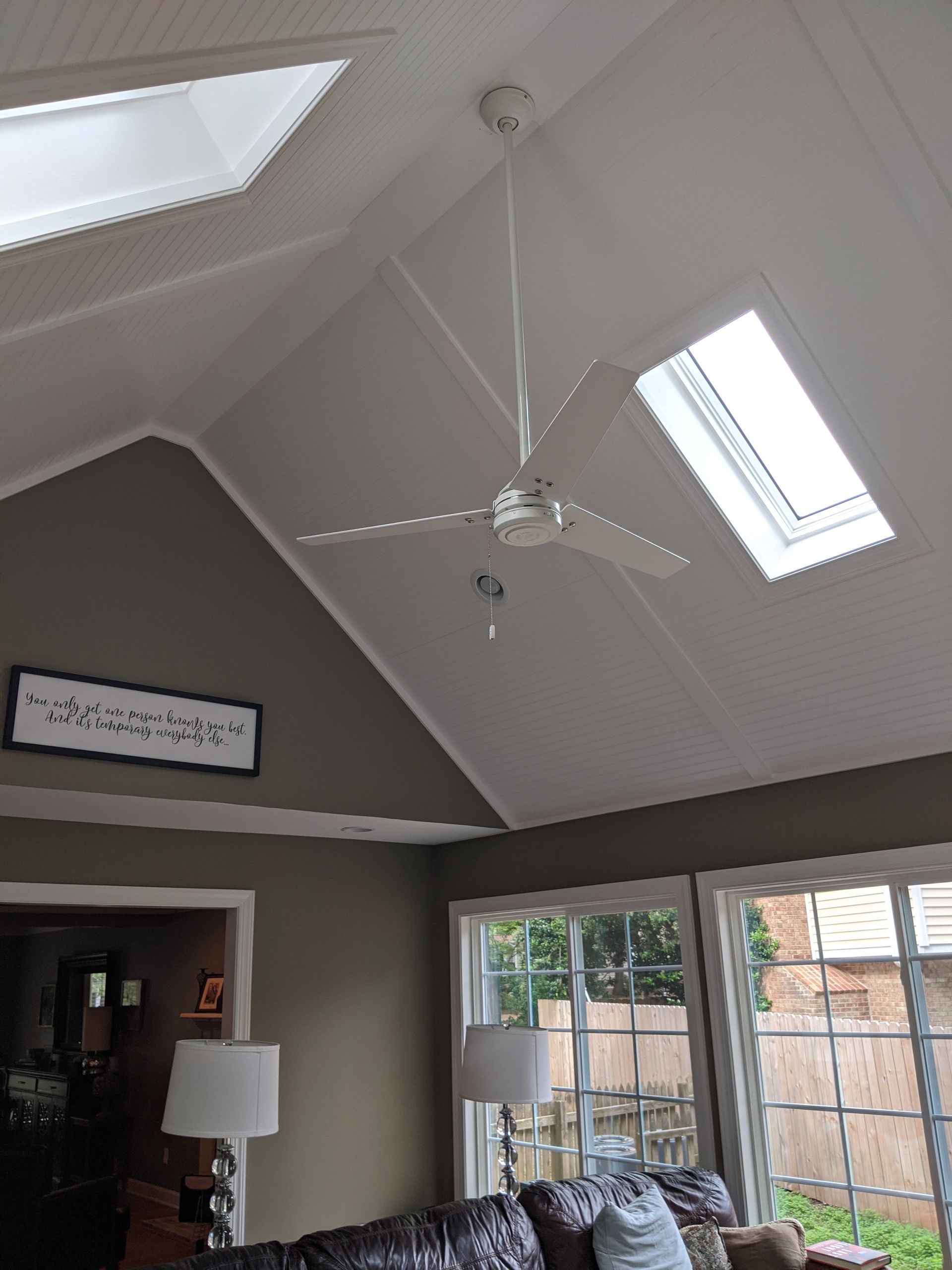 A white ceiling fan hangs in a room with a vaulted white ceiling, two skylights, and taupe walls above a seating area.