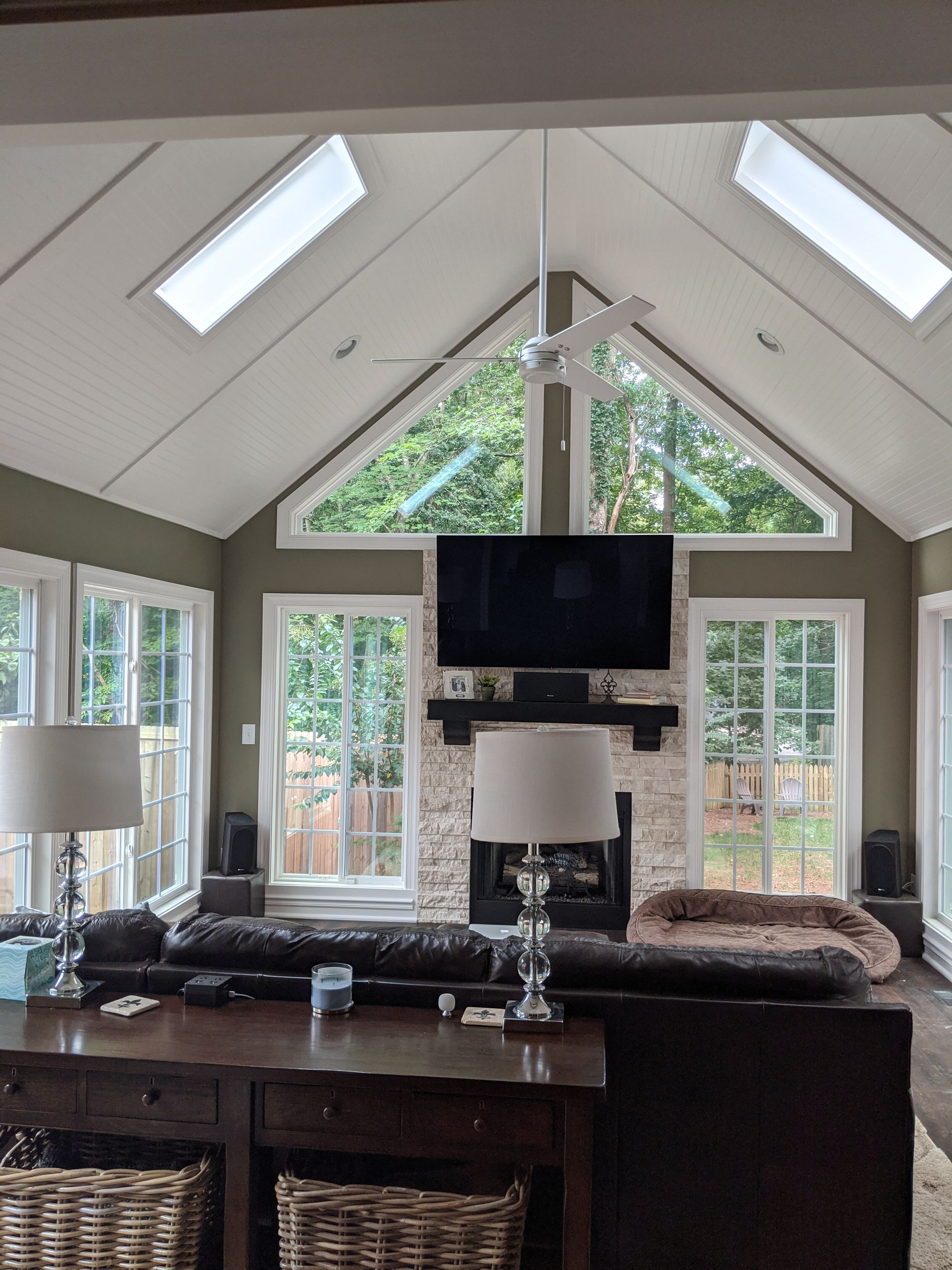 A cozy living room with a stone fireplace, large windows, vaulted ceiling with skylights, and a plush brown sofa.