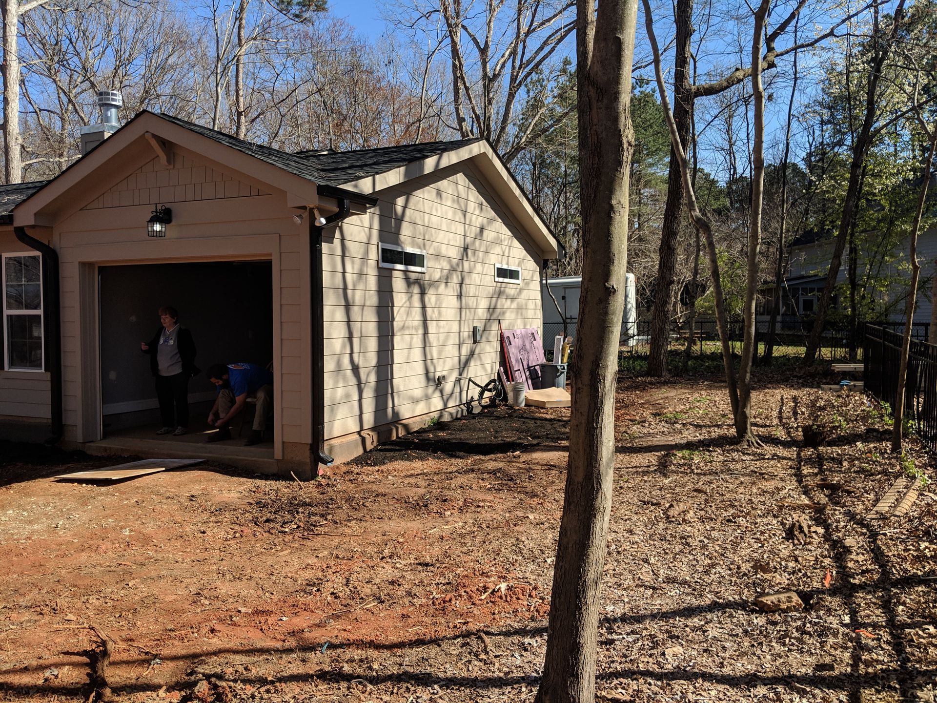 A garage with a beige stucco exterior and a dark roof, surrounded by trees and a dirt yard with two people inside.