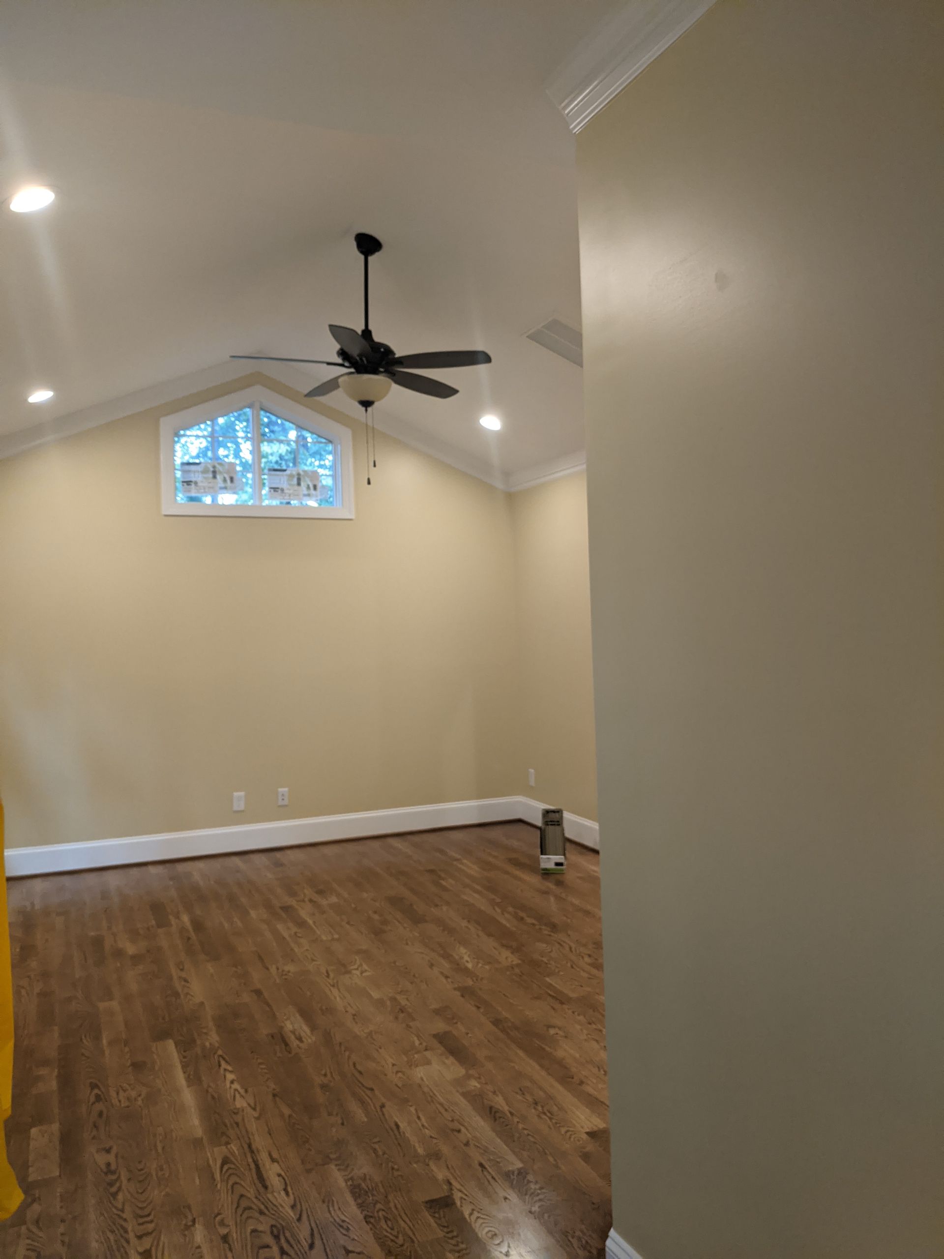 An empty room with beige walls, vaulted ceilings, a dark wood floor, a ceiling fan, and a transom window.