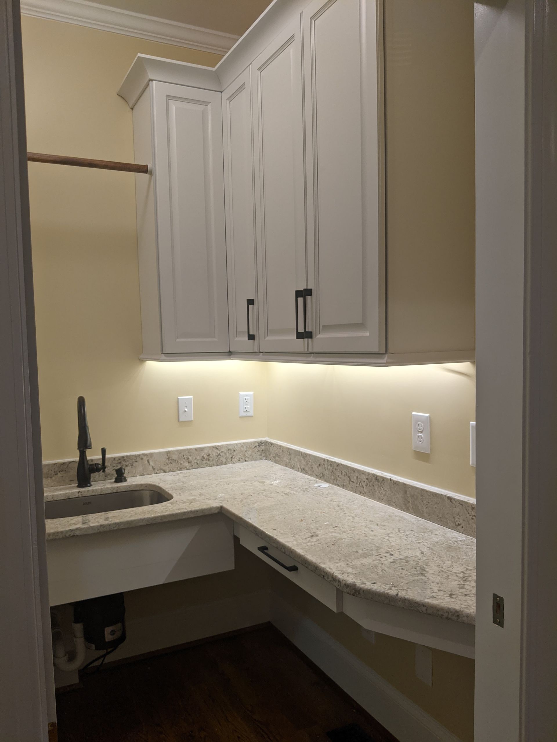 Laundry room corner with light-colored cabinets, granite countertops, a dark faucet, and under-cabinet lighting.