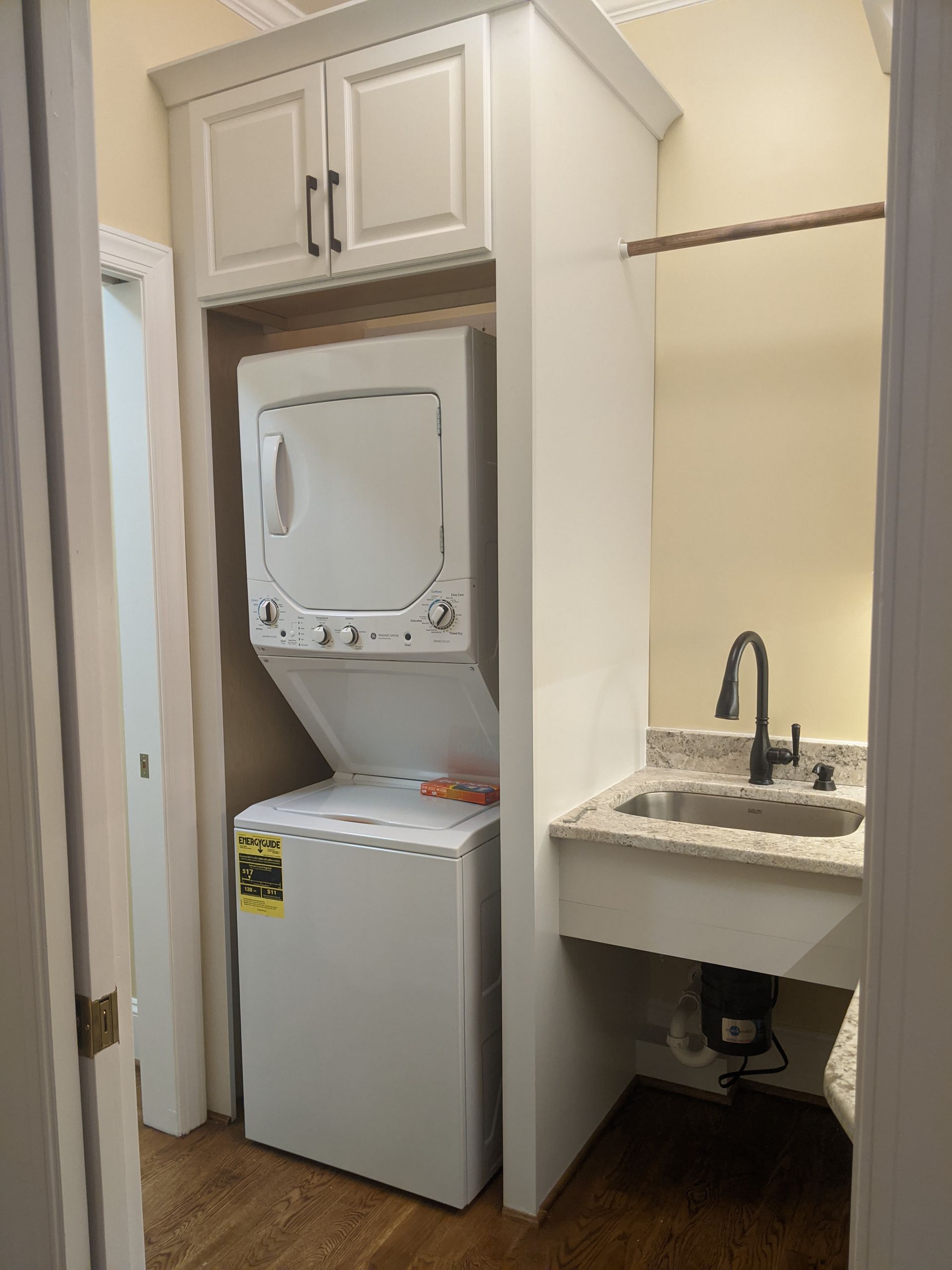 A white laundry nook features a stacked washer-dryer unit next to a small utility sink with a black faucet on a countertop.