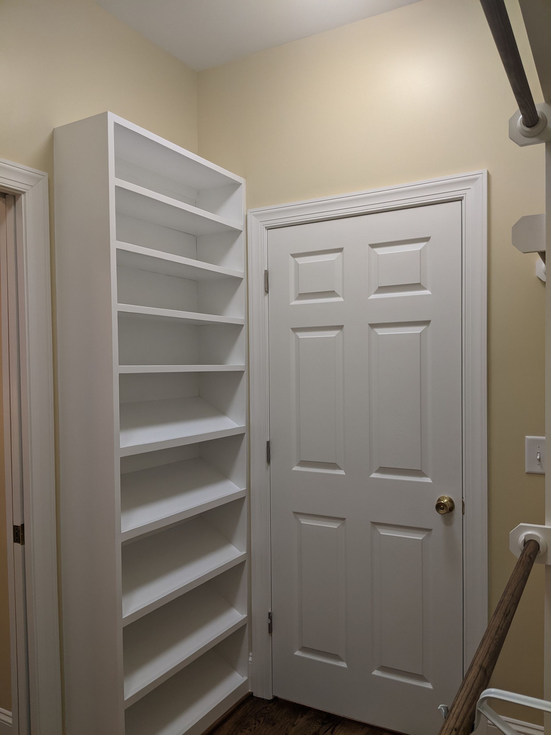 A white floor-to-ceiling shelving unit sits next to a closed white six-panel door in a room with tan walls.