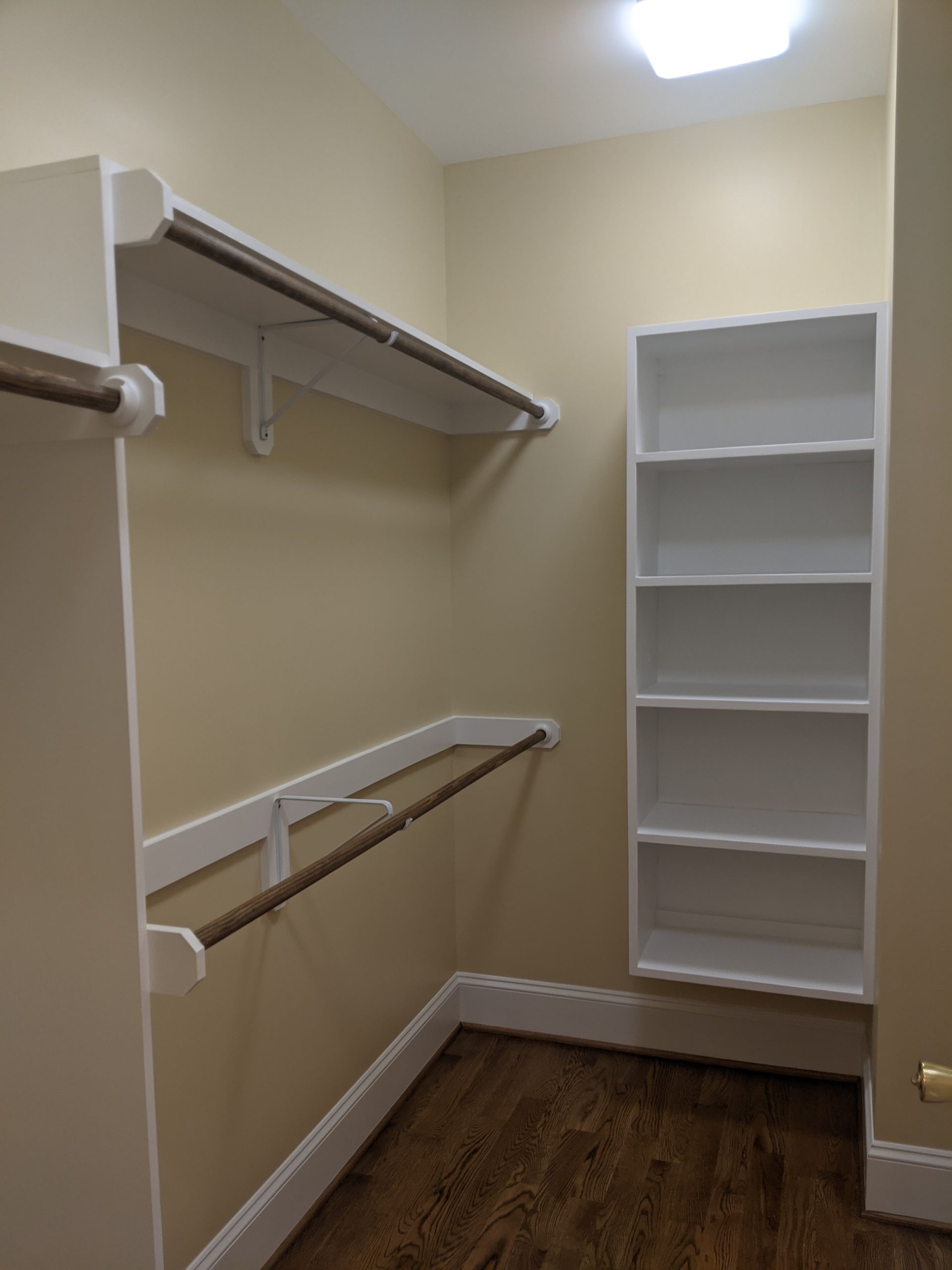 A walk-in closet featuring white wooden shelving units, two hanging rods, beige walls, and dark hardwood flooring.