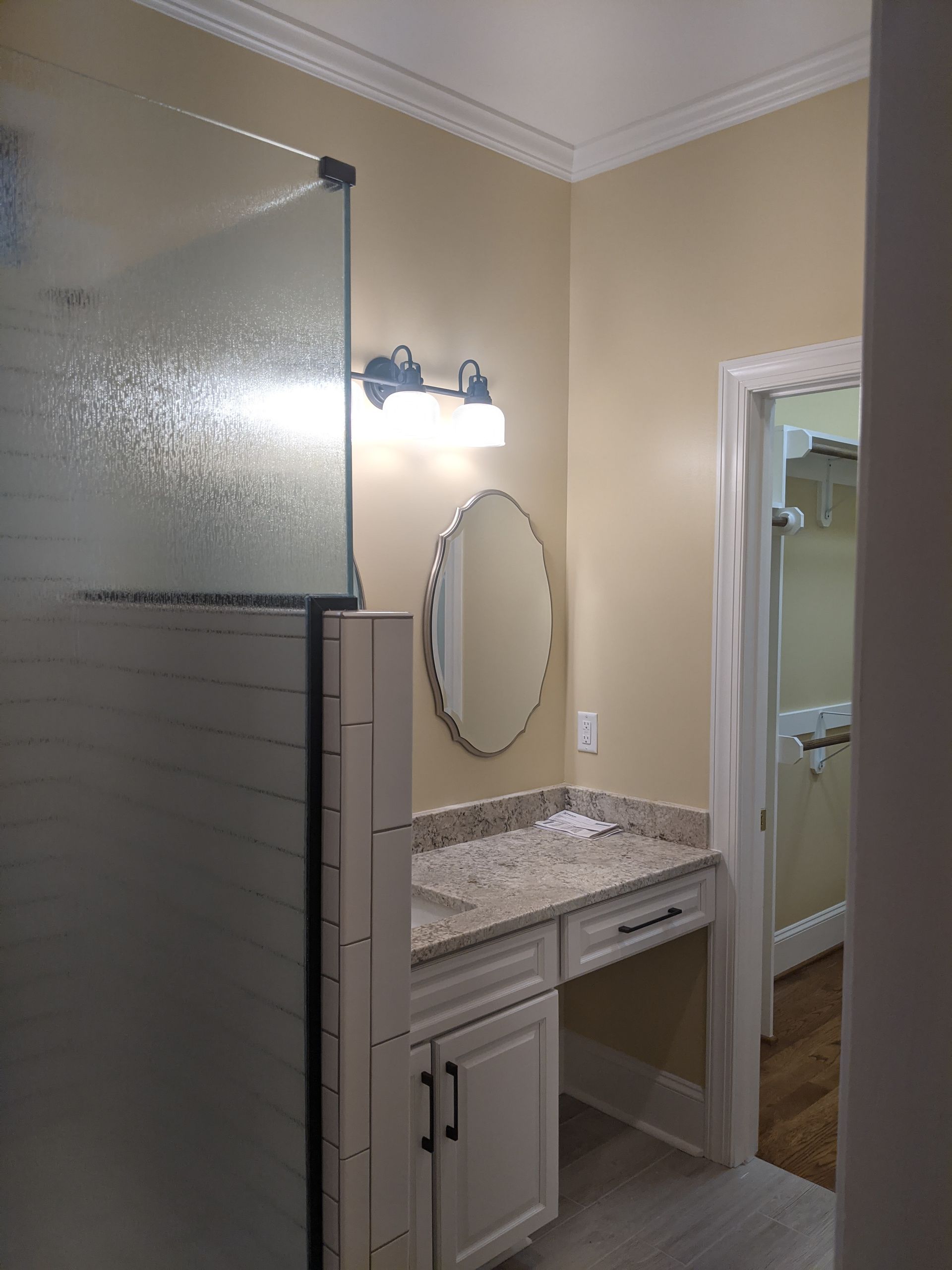 A bathroom vanity with a granite countertop, oval mirror, wall lighting, and a frosted glass shower door.