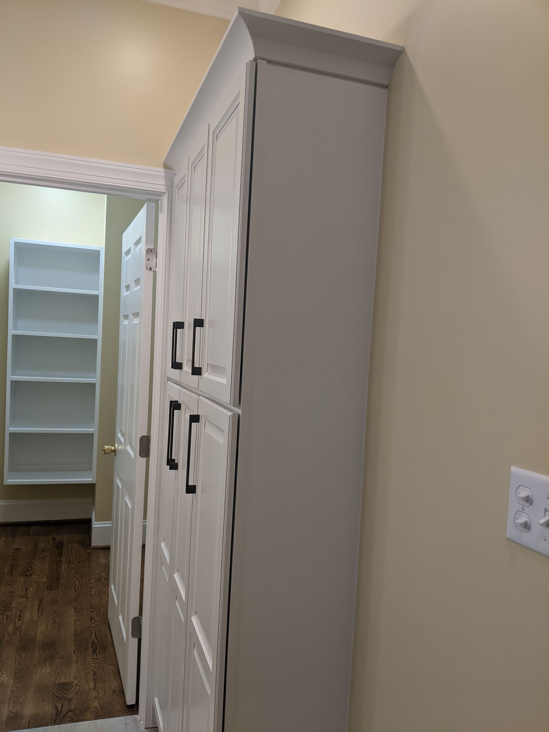 Tall white pantry cabinet with black handles in a room adjacent to a doorway and a built-in shelving unit.