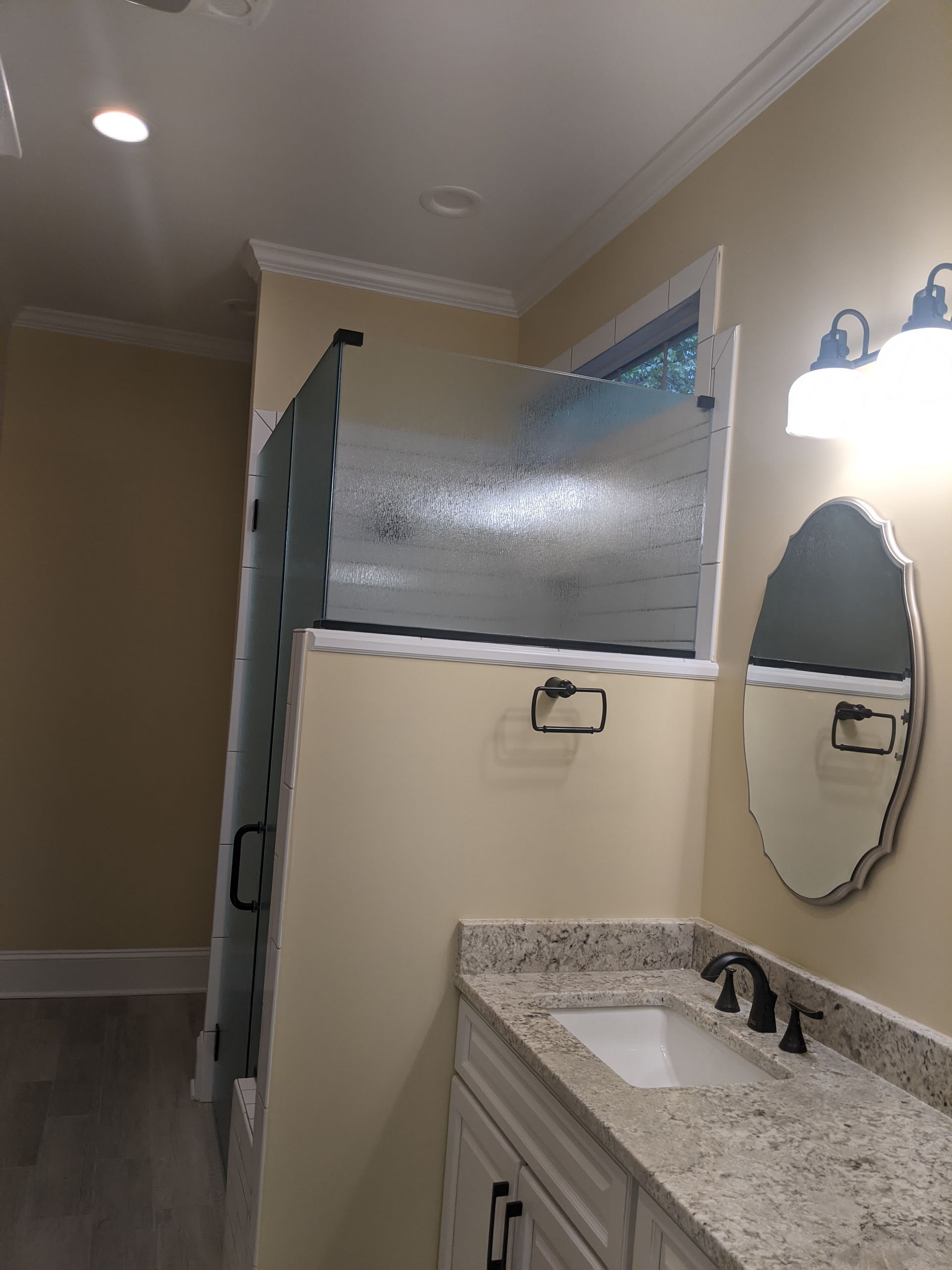 A bathroom with a granite vanity, oval mirror, wall sconces, and a shower with textured glass panels.