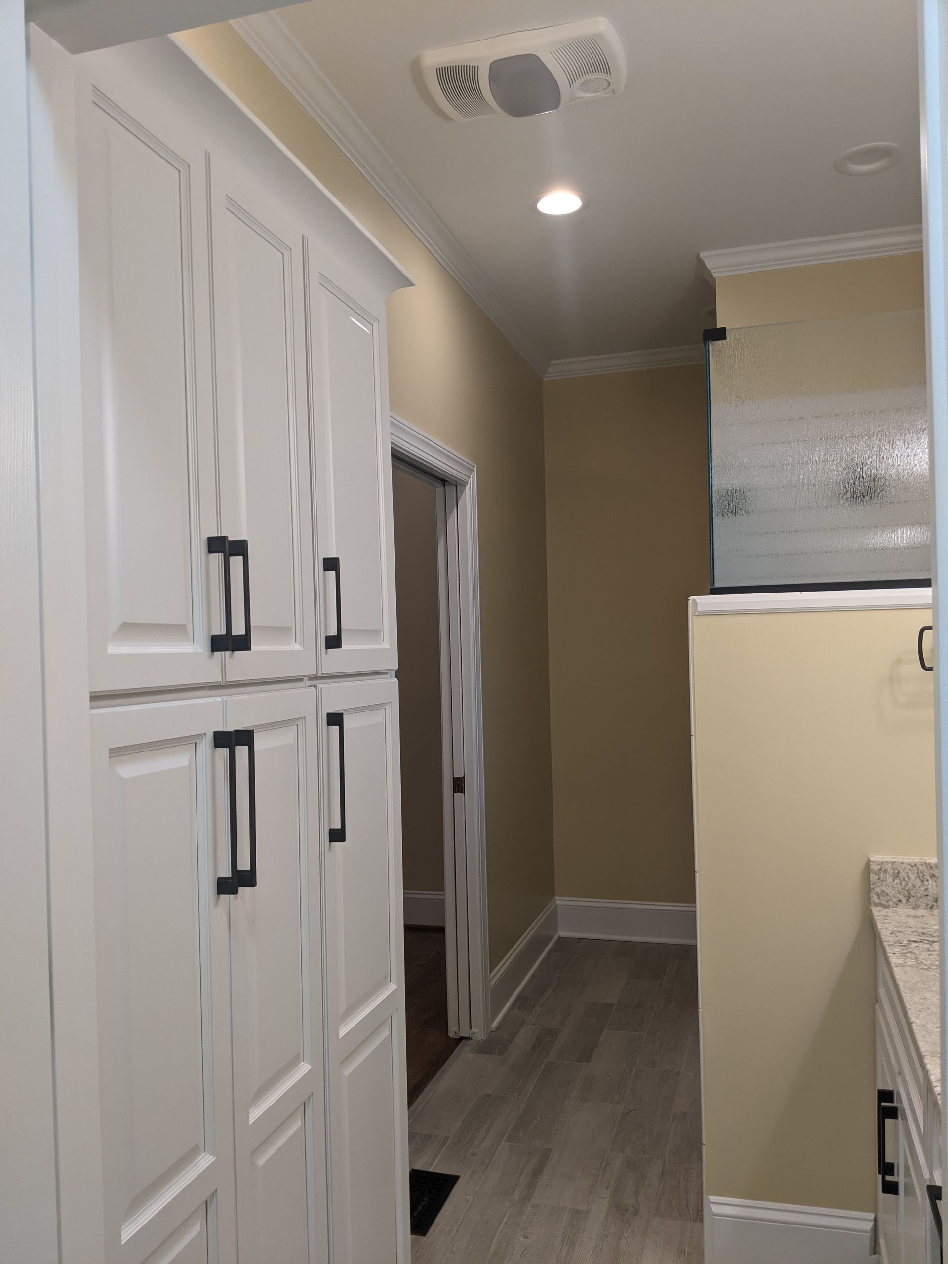 A hallway featuring tall white cabinets with black handles, light wood flooring, tan walls, and a recessed ceiling light.