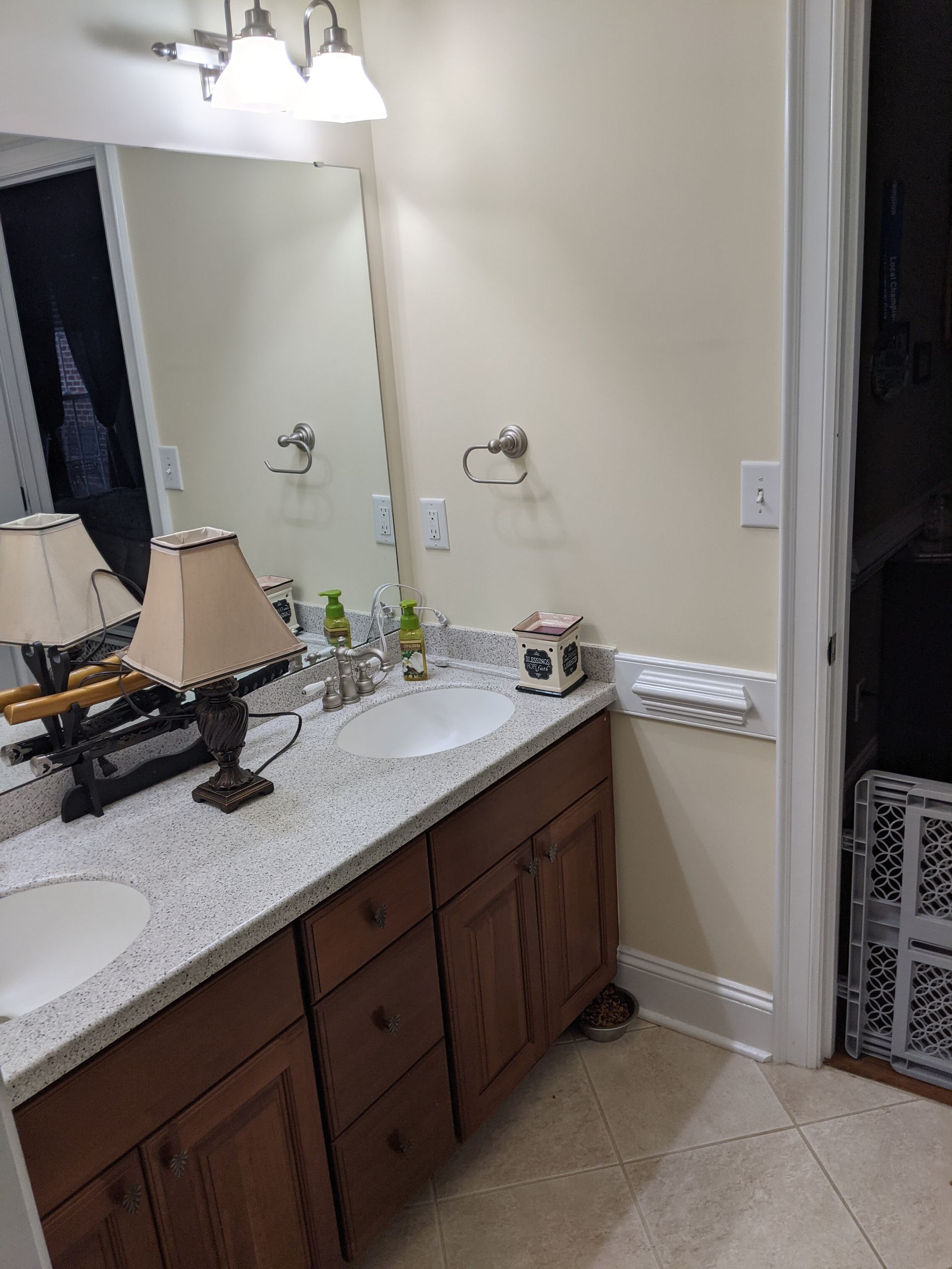 Bathroom vanity with a white speckled countertop, two sinks, dark wood cabinets, and two lamp accents on the counter.