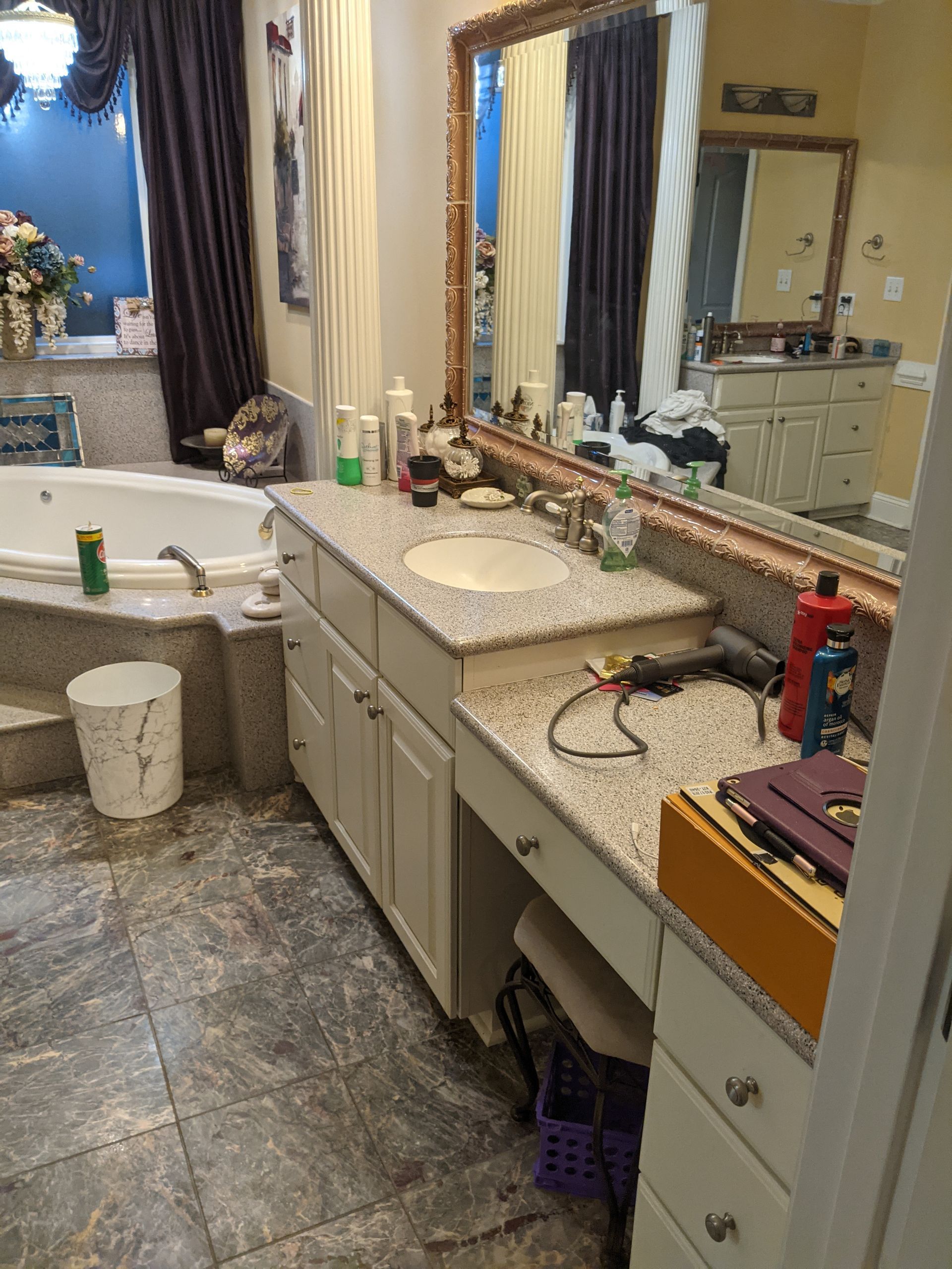A bathroom featuring a granite-topped double vanity with a mirror, a soaking tub, and dark curtains.
