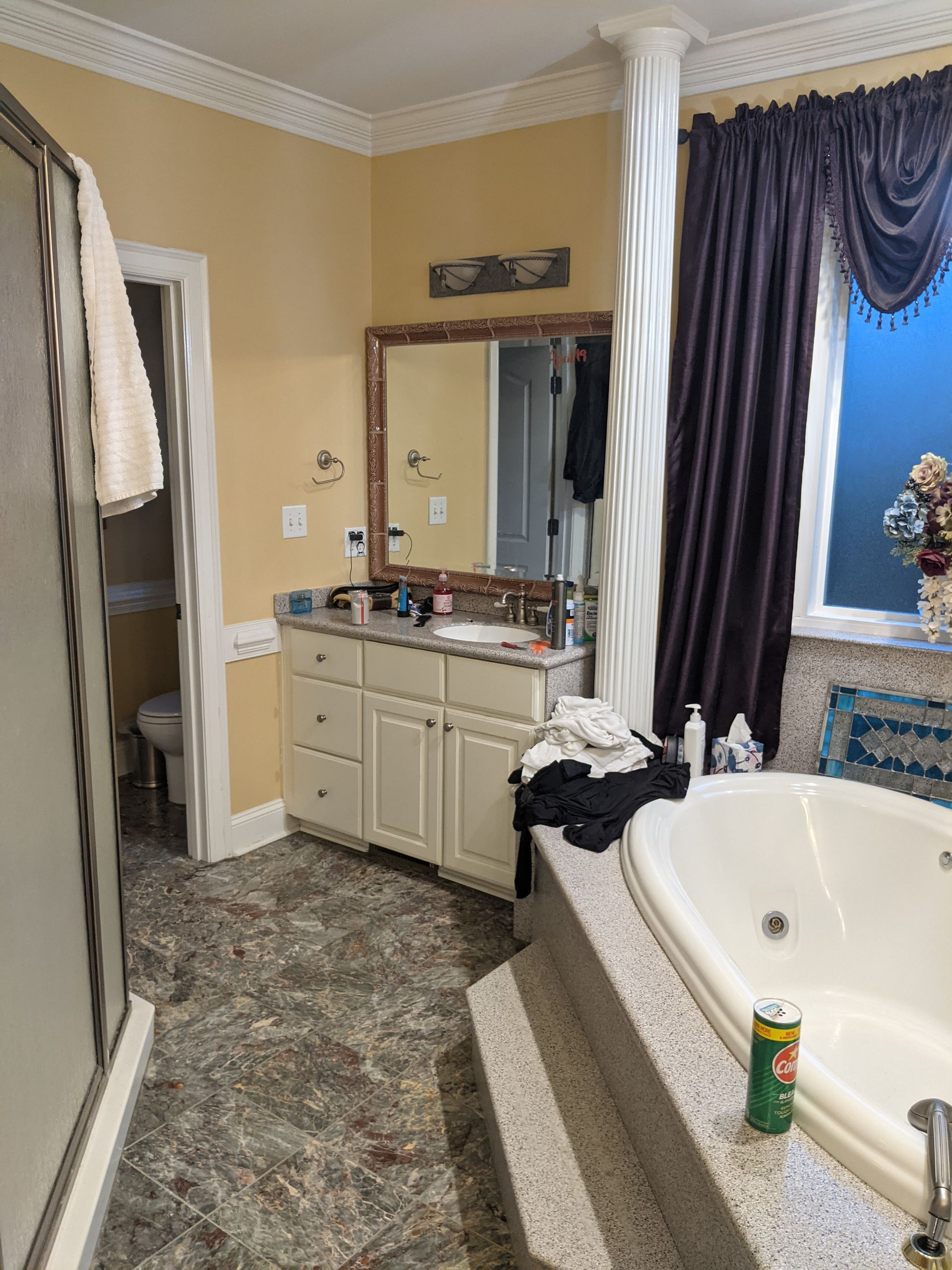 A bathroom featuring a cream vanity, a jetted tub with a stone deck, dark curtains, and a textured white pillar.
