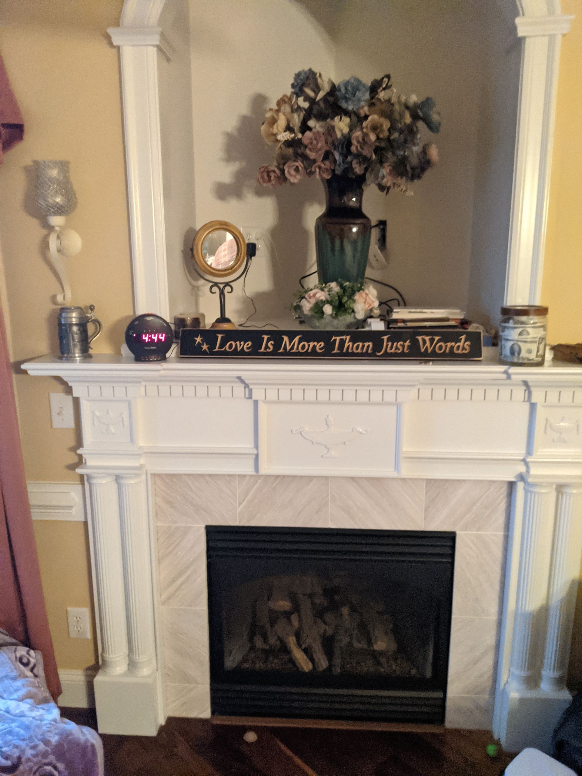 A decorative white fireplace mantle displays a vase of flowers, a small mirror, and a sign reading 
