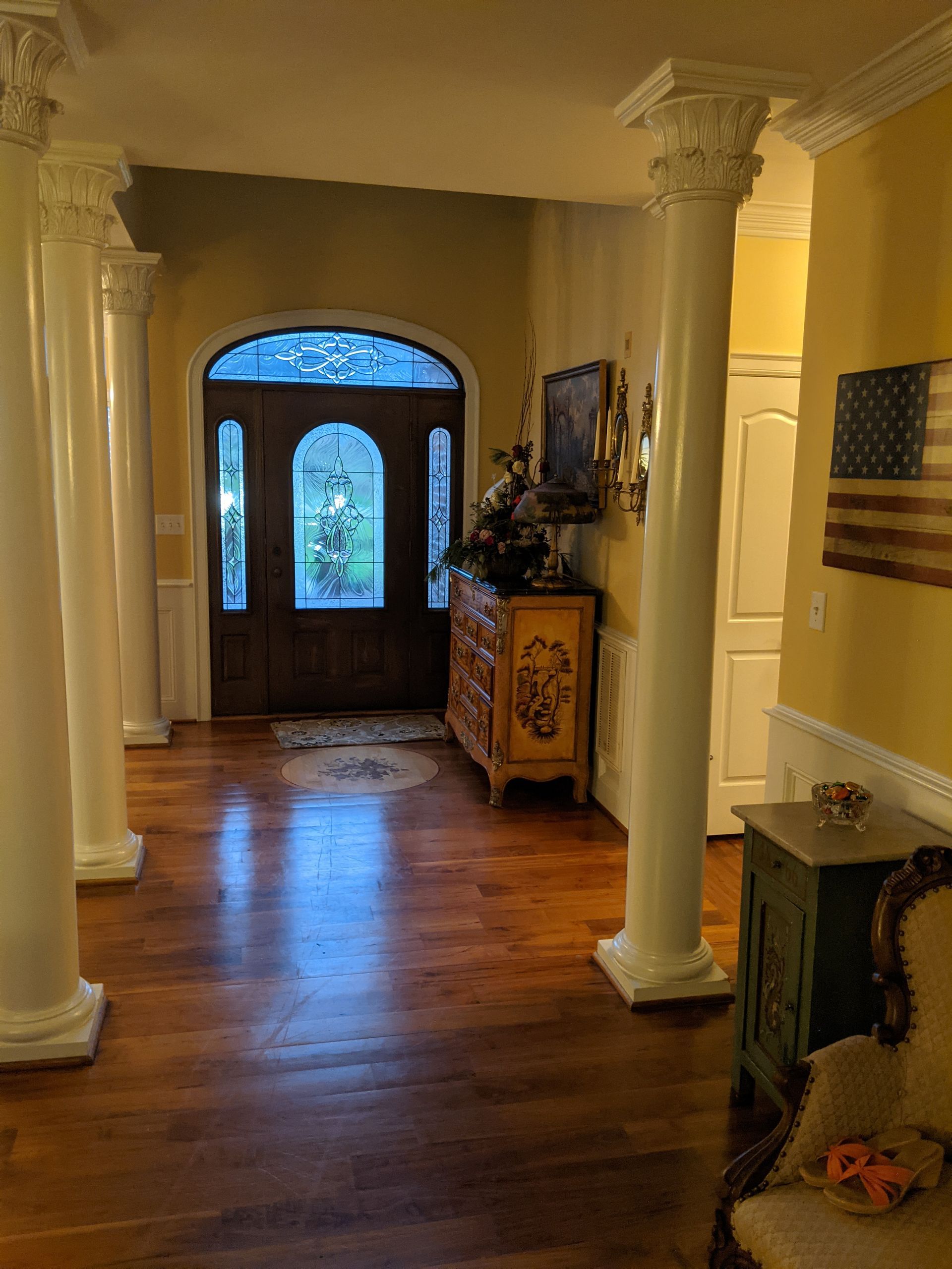 A foyer with wooden floors, tall white pillars, an arched front door, a decorative cabinet, and a wooden flag on the wall.