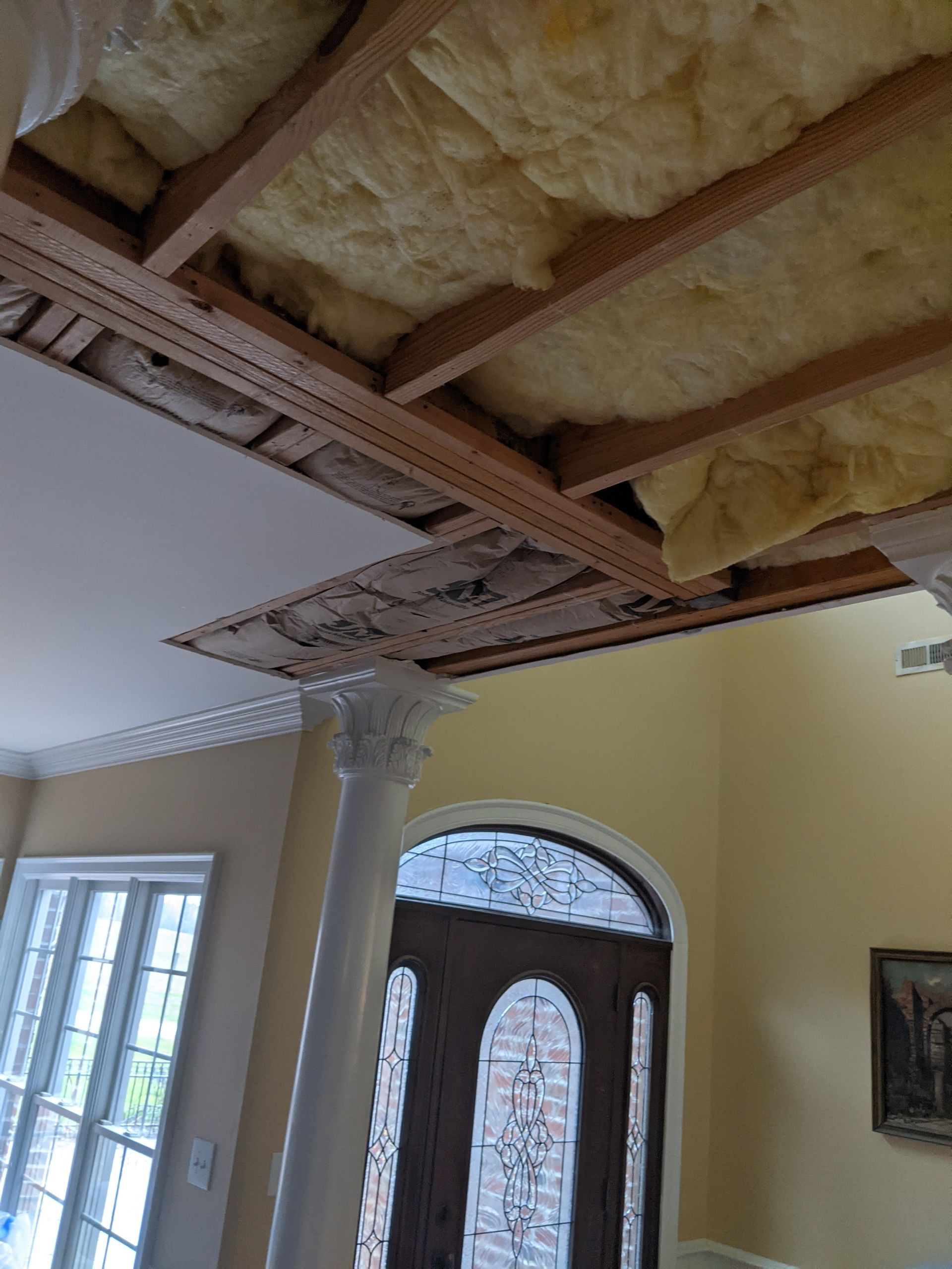 A room with a partially open ceiling revealing wooden joists and yellow insulation, with a white column in the foreground.