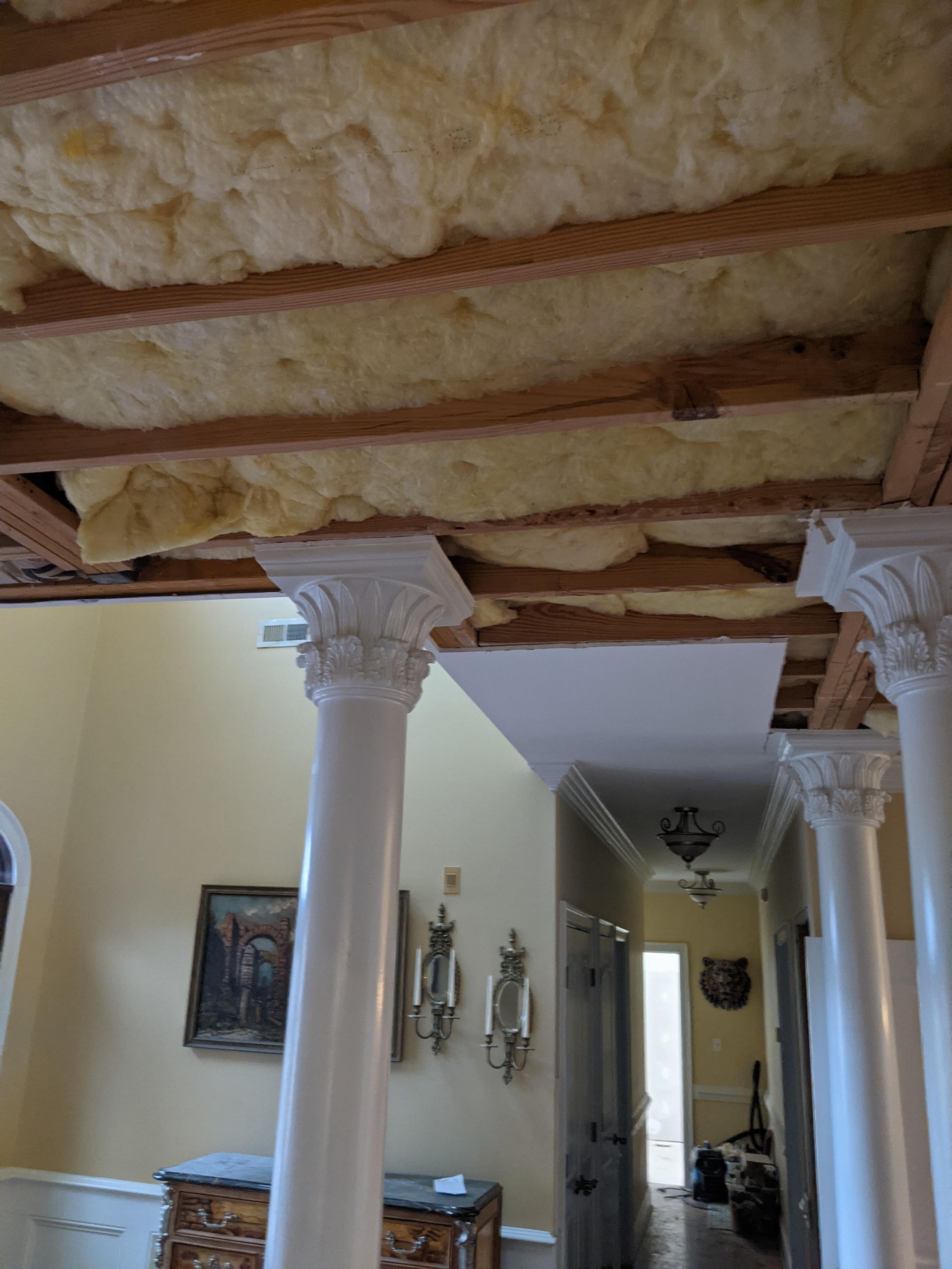The ceiling in a residential hallway is exposed, revealing wooden joists and yellow fiberglass insulation above pillars.