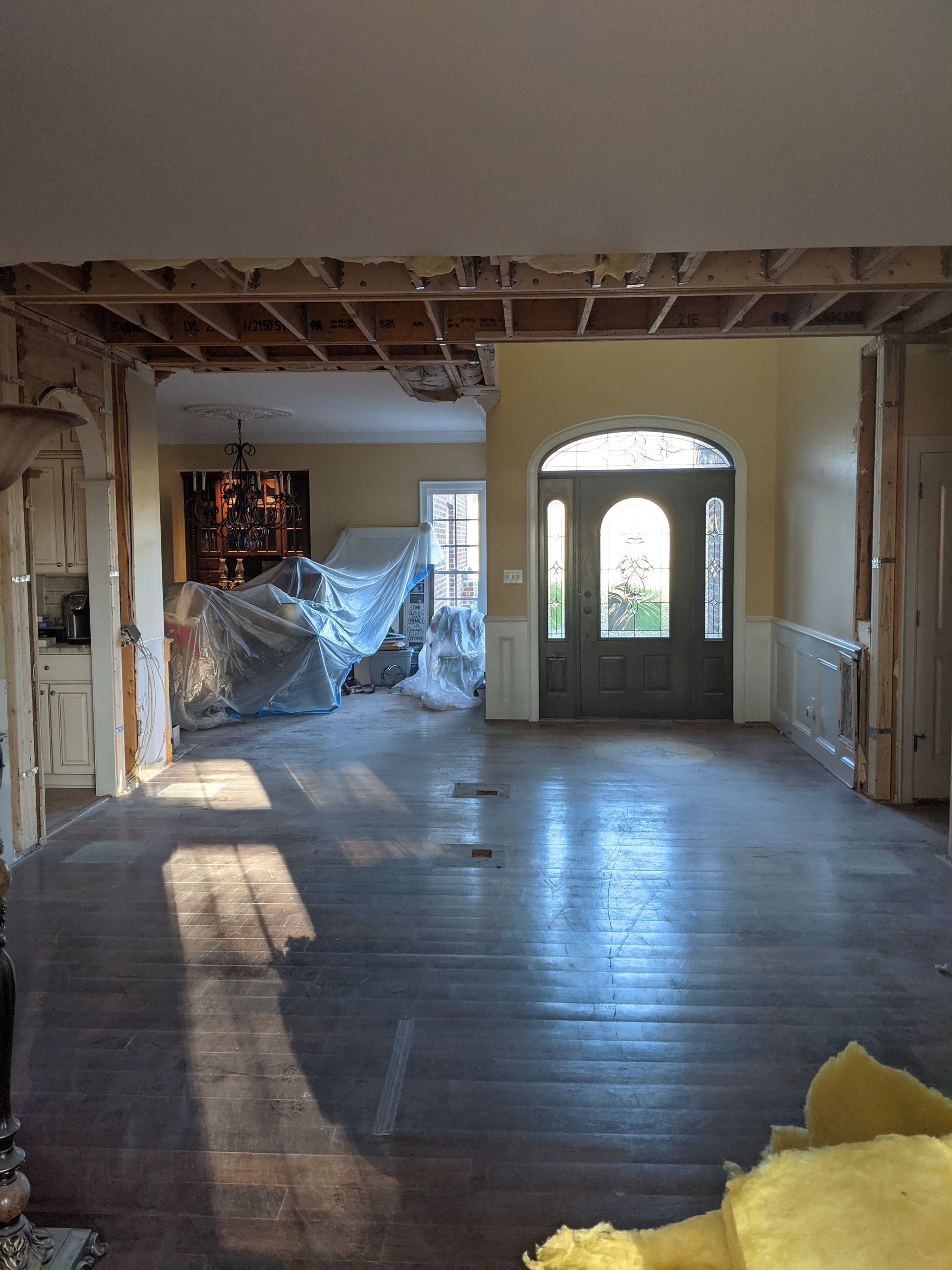 An interior view of a room under construction, showing wooden ceiling beams, bare floors, and a front entry door.