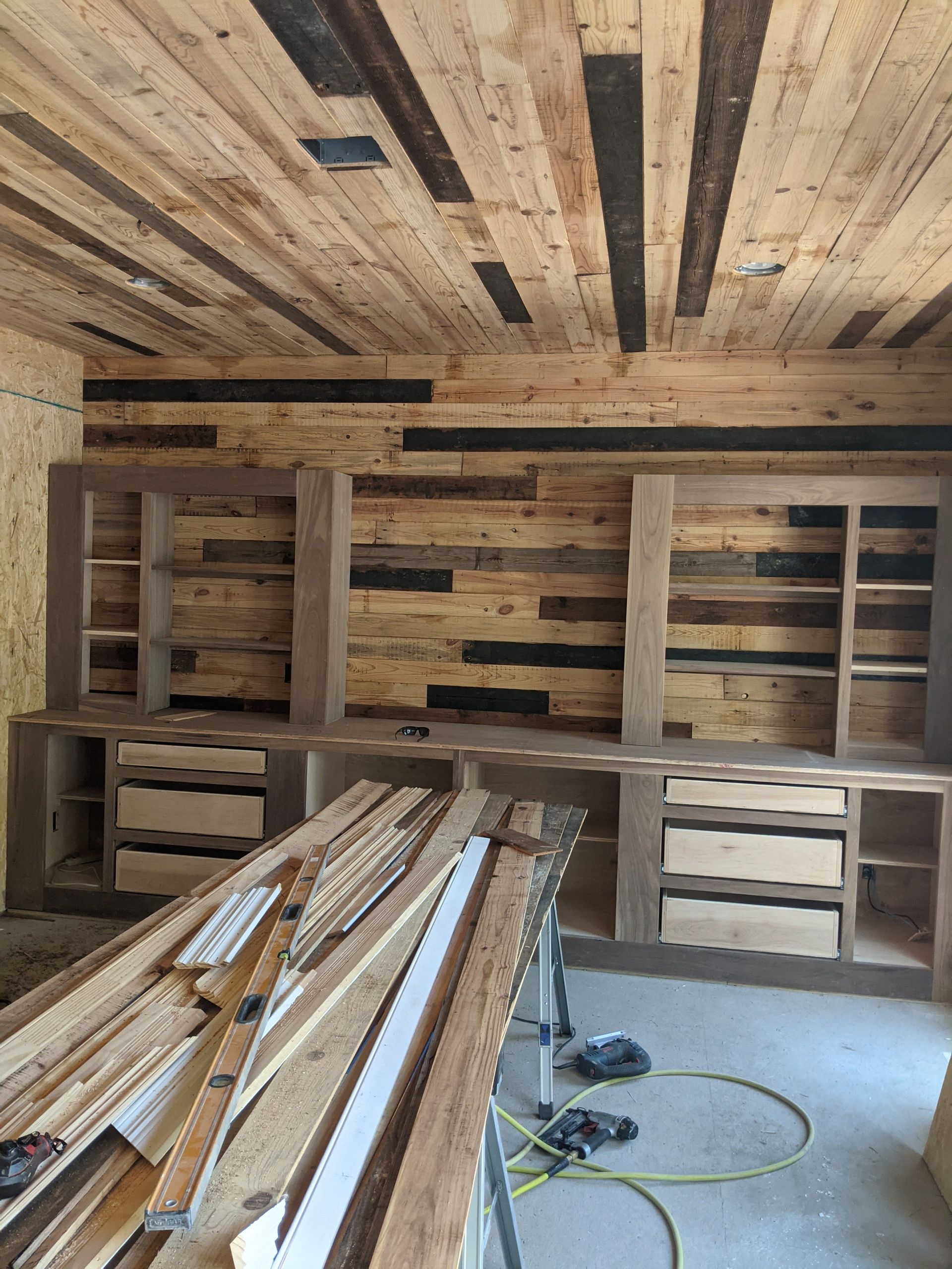 A room under construction with rustic wood-paneled walls and ceiling, featuring built-in wooden shelving and desk units.