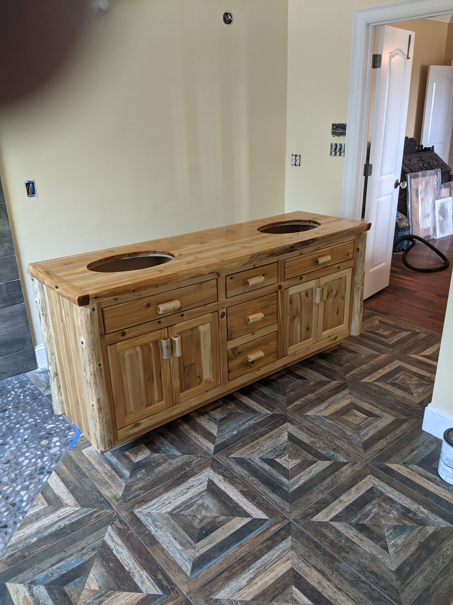 A rustic, light-wood double vanity cabinet with two sink cutouts, positioned in a room with a patterned tile floor.