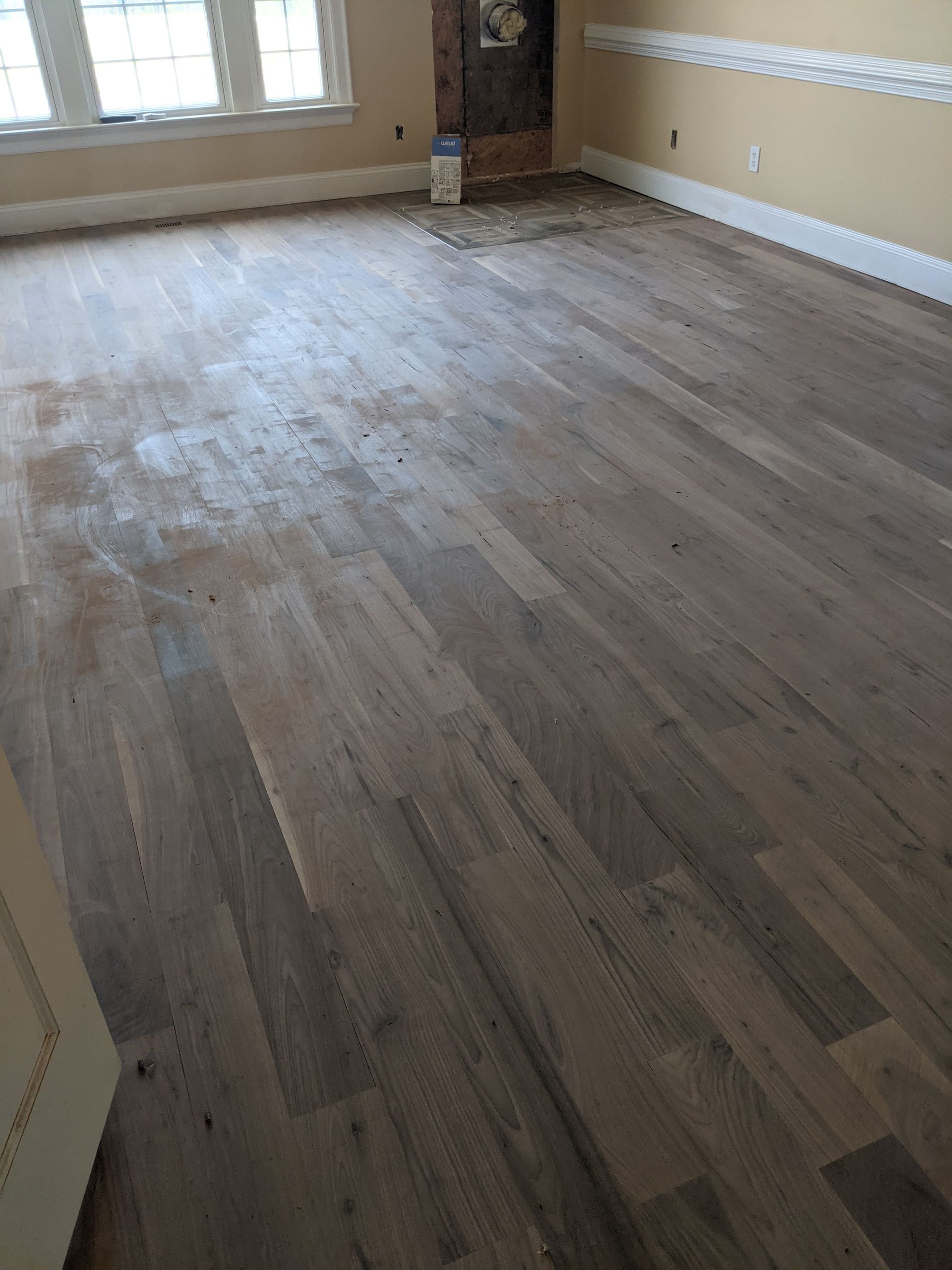 Hardwood floor in a room, partially sanded with some unfinished patches, light beige walls, and white trim.