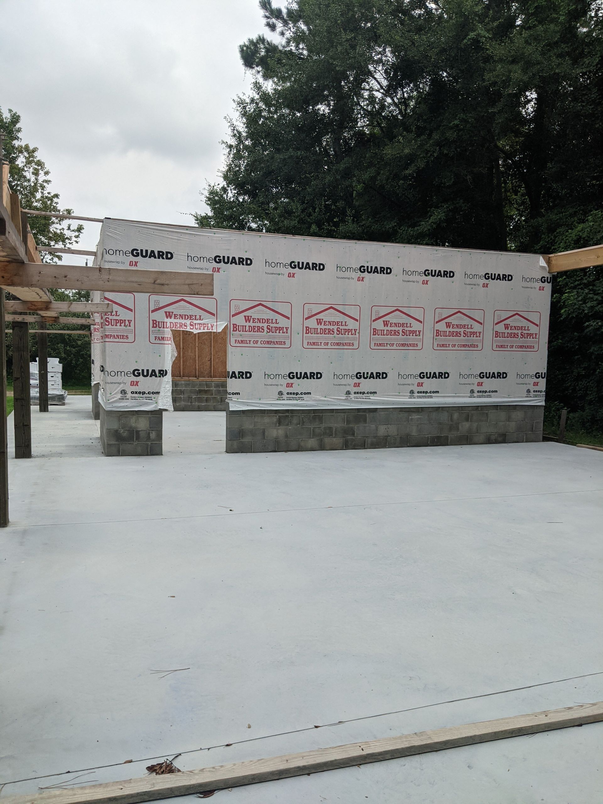 A partially constructed building with concrete foundation walls and wood framing wrapped in white moisture barrier sheeting.
