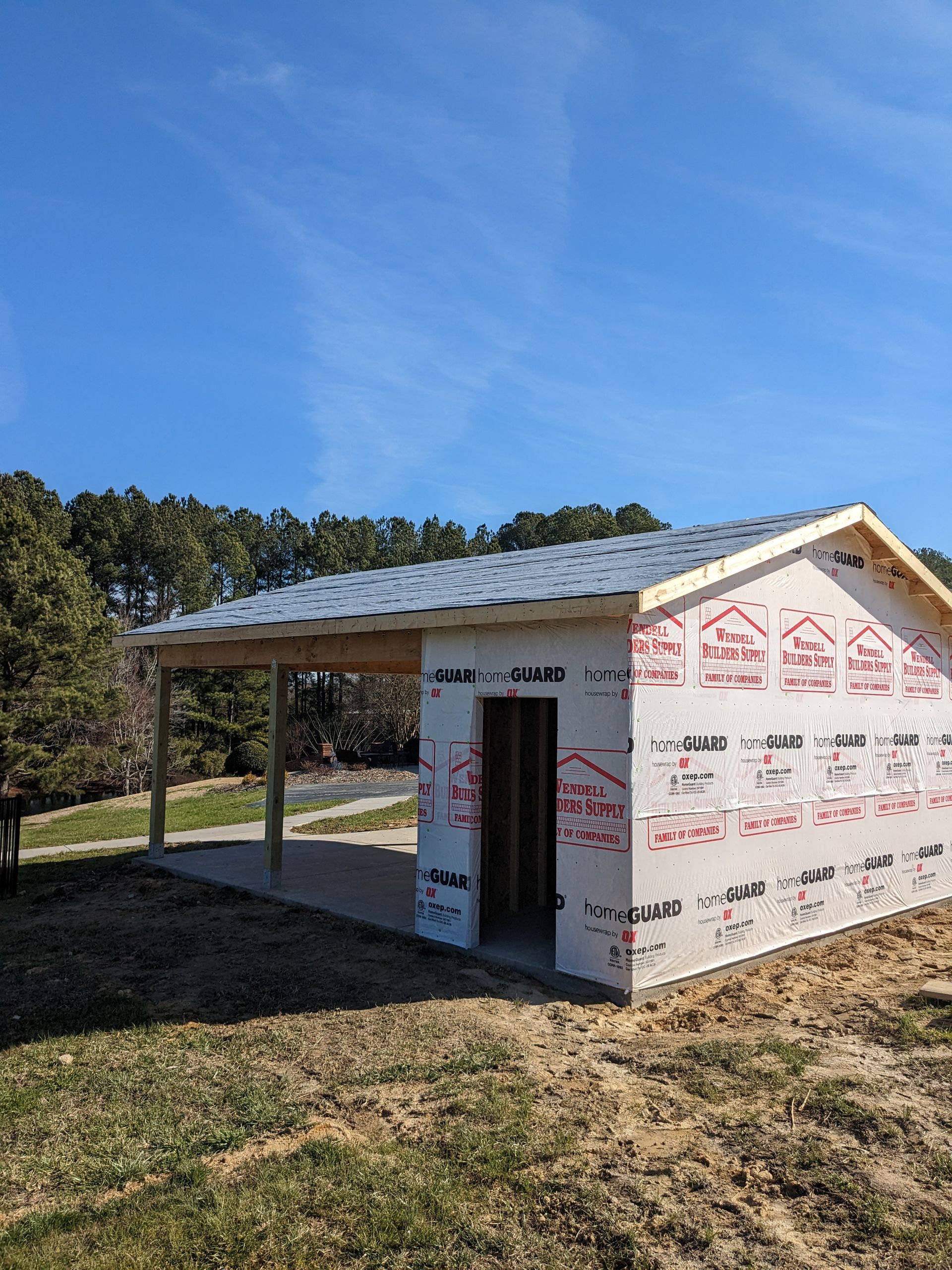 An unfinished shed with a covered porch and white foam-board siding sits in a grassy yard under a clear blue sky.