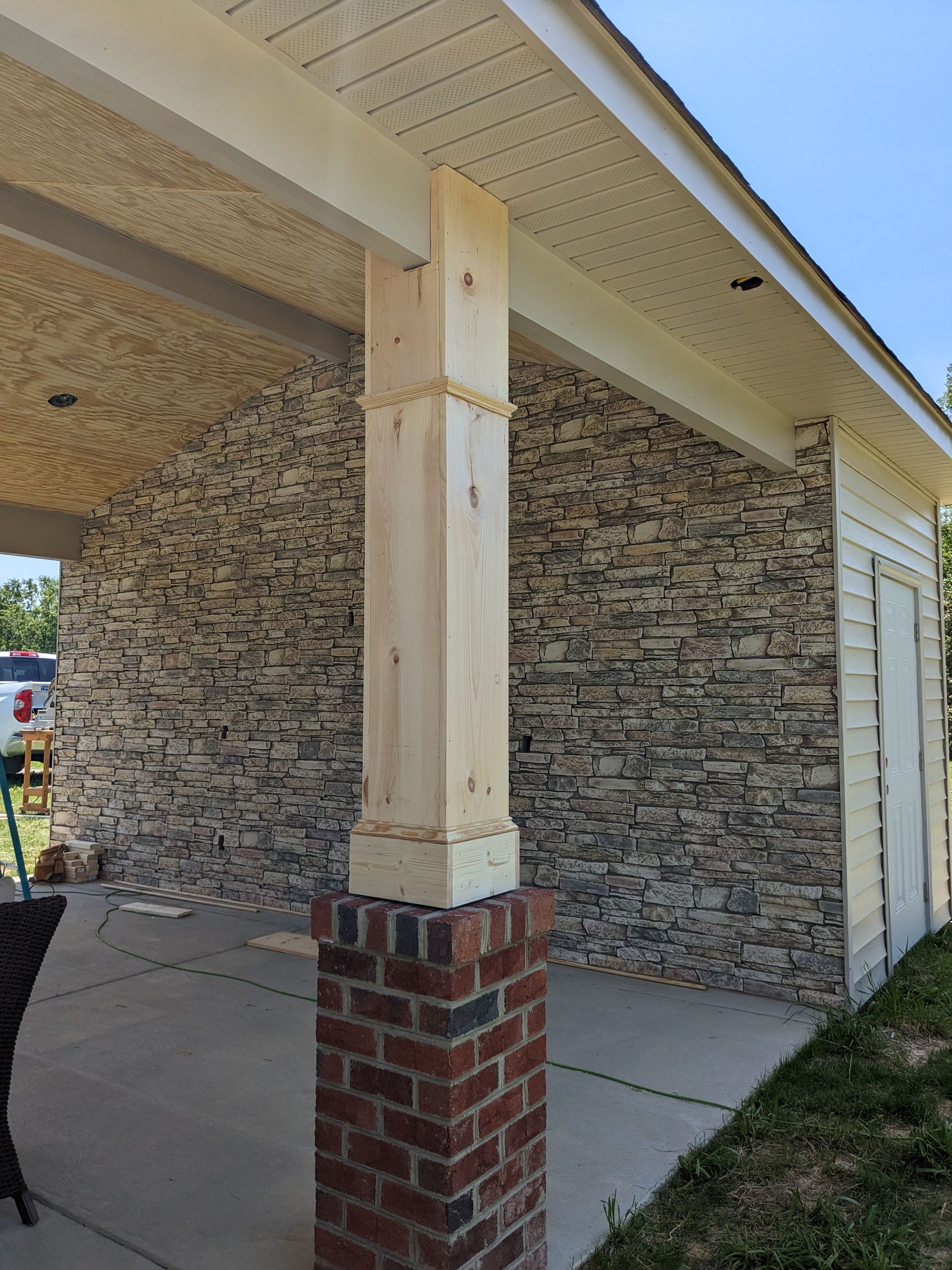 A brick pillar topped with a wooden post supports the porch roof of a building with a patterned, stone-look wall.