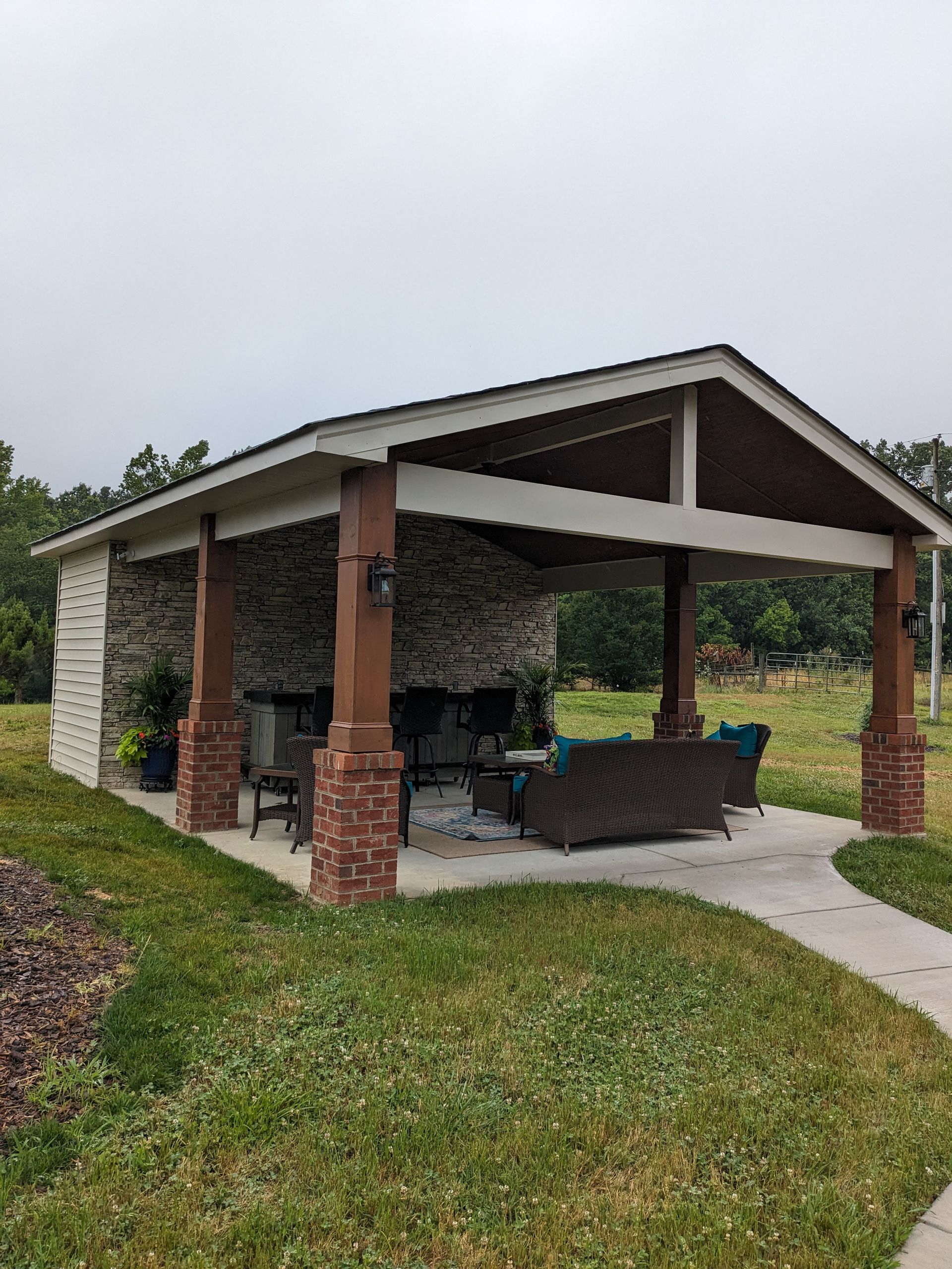 Outdoor pavilion with stone walls, wooden pillars, and patio furniture on a concrete slab in a grassy yard.