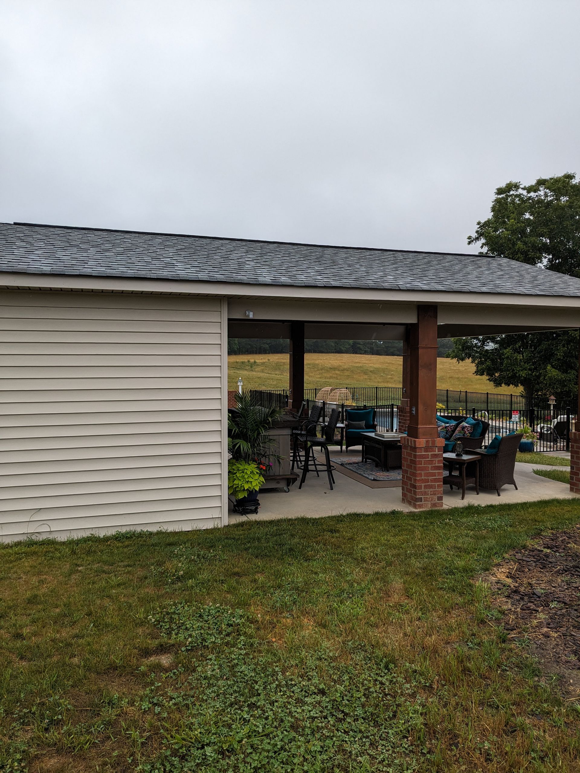 A tan, vinyl-sided patio structure with a dark shingled roof, featuring a brick-columned covered area with outdoor seating.