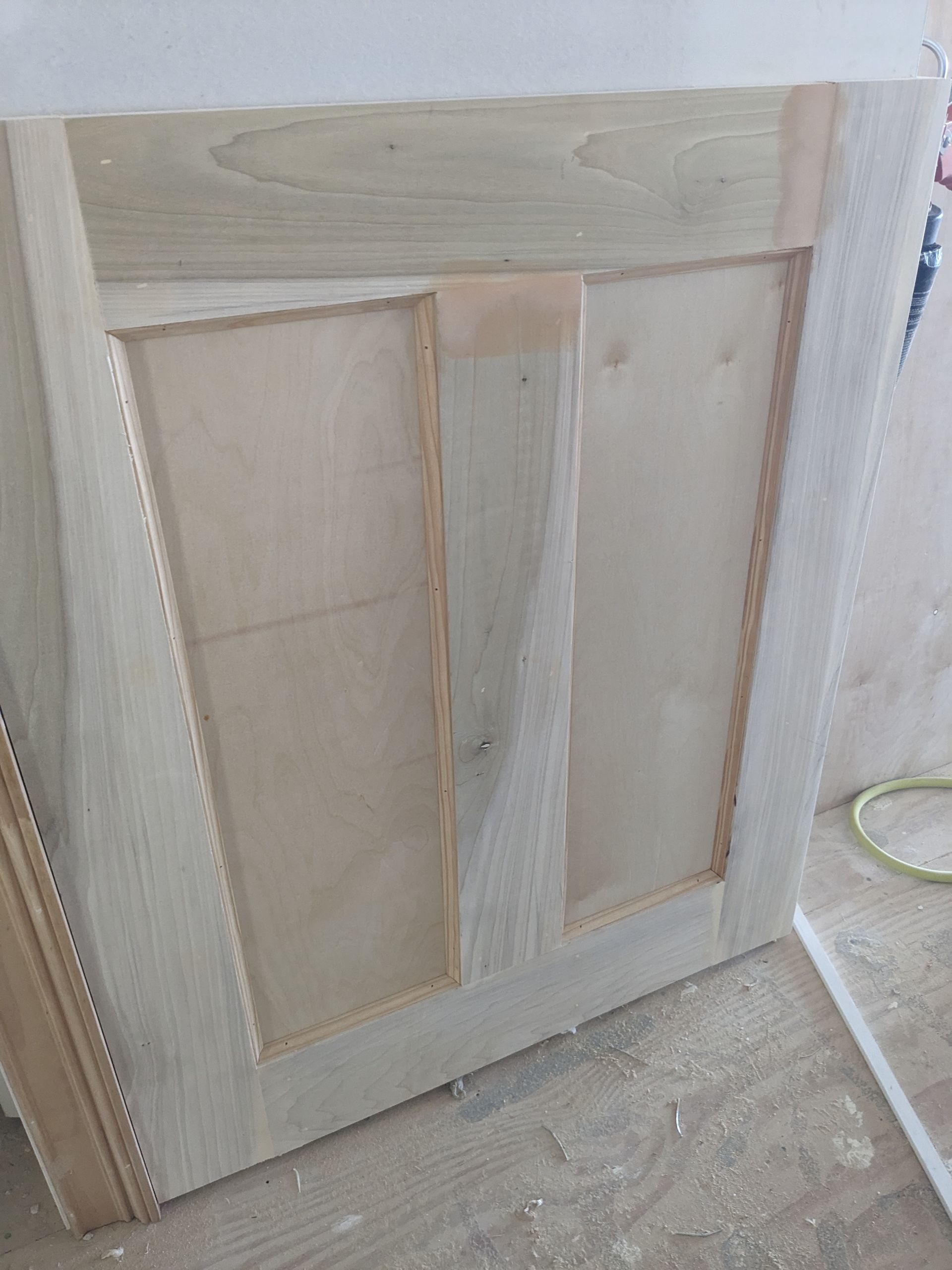 A light-colored wooden cabinet door being built, showing a frame and panels with visible joinery and light wood grain.