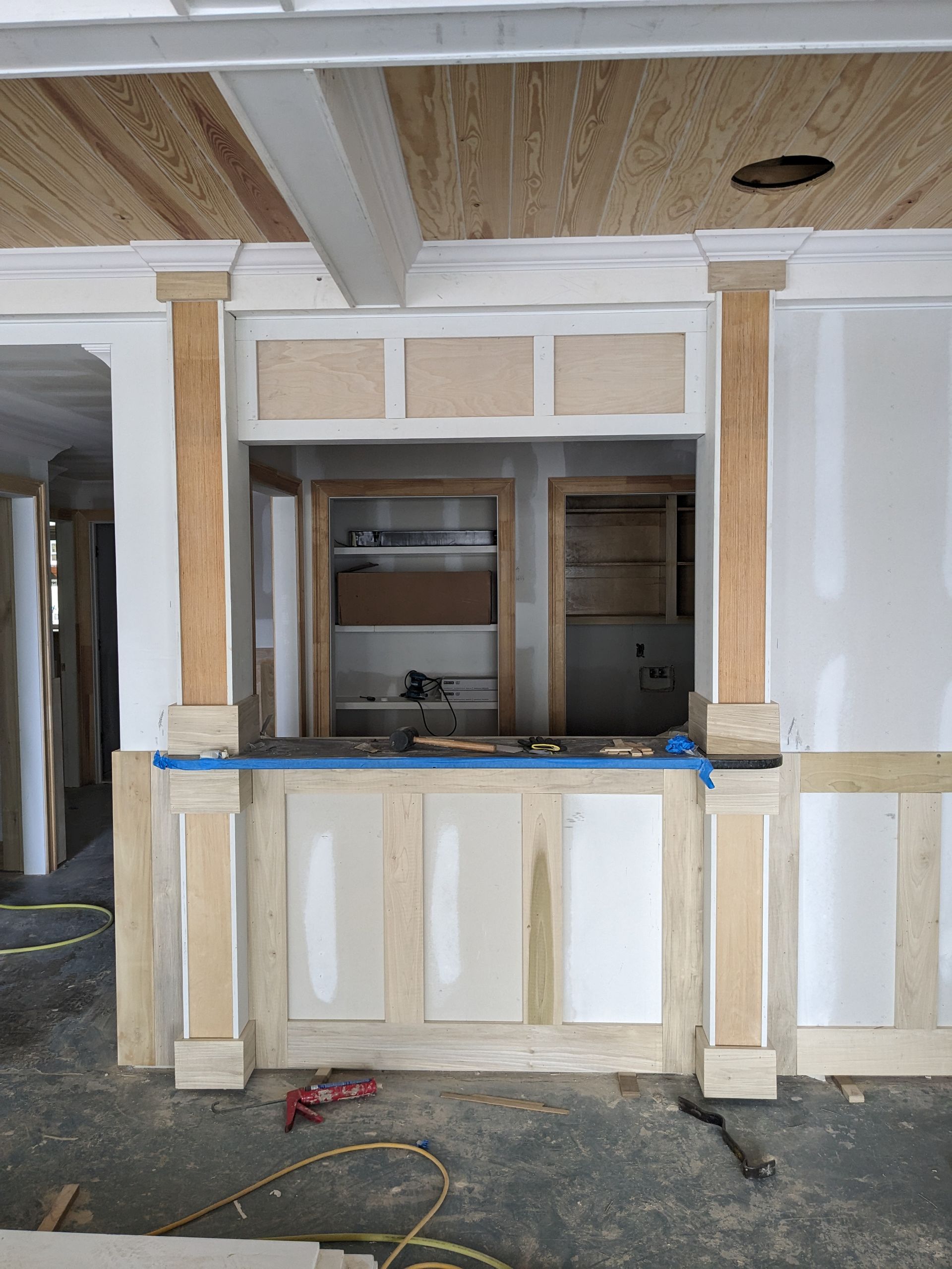 Unfinished basement bar framing featuring wood trim pillars, paneling, and an open pass-through window with storage behind.