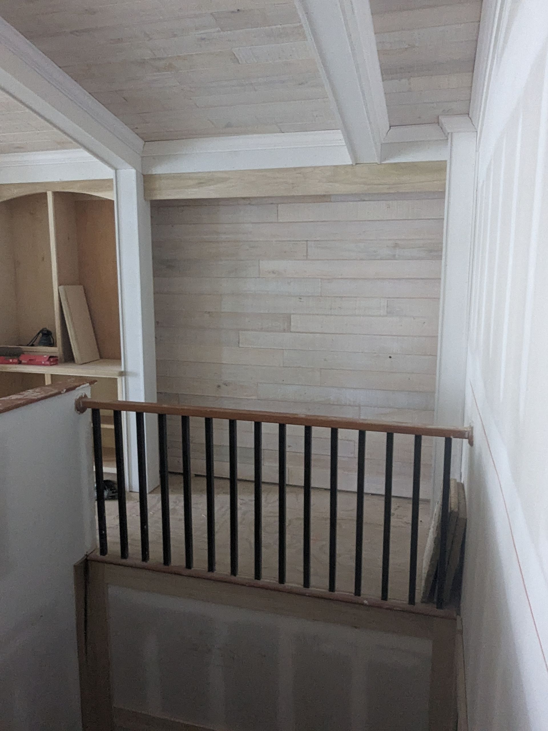 A residential loft area featuring a light-toned wood plank wall, white-painted ceiling beams, and a black metal railing.