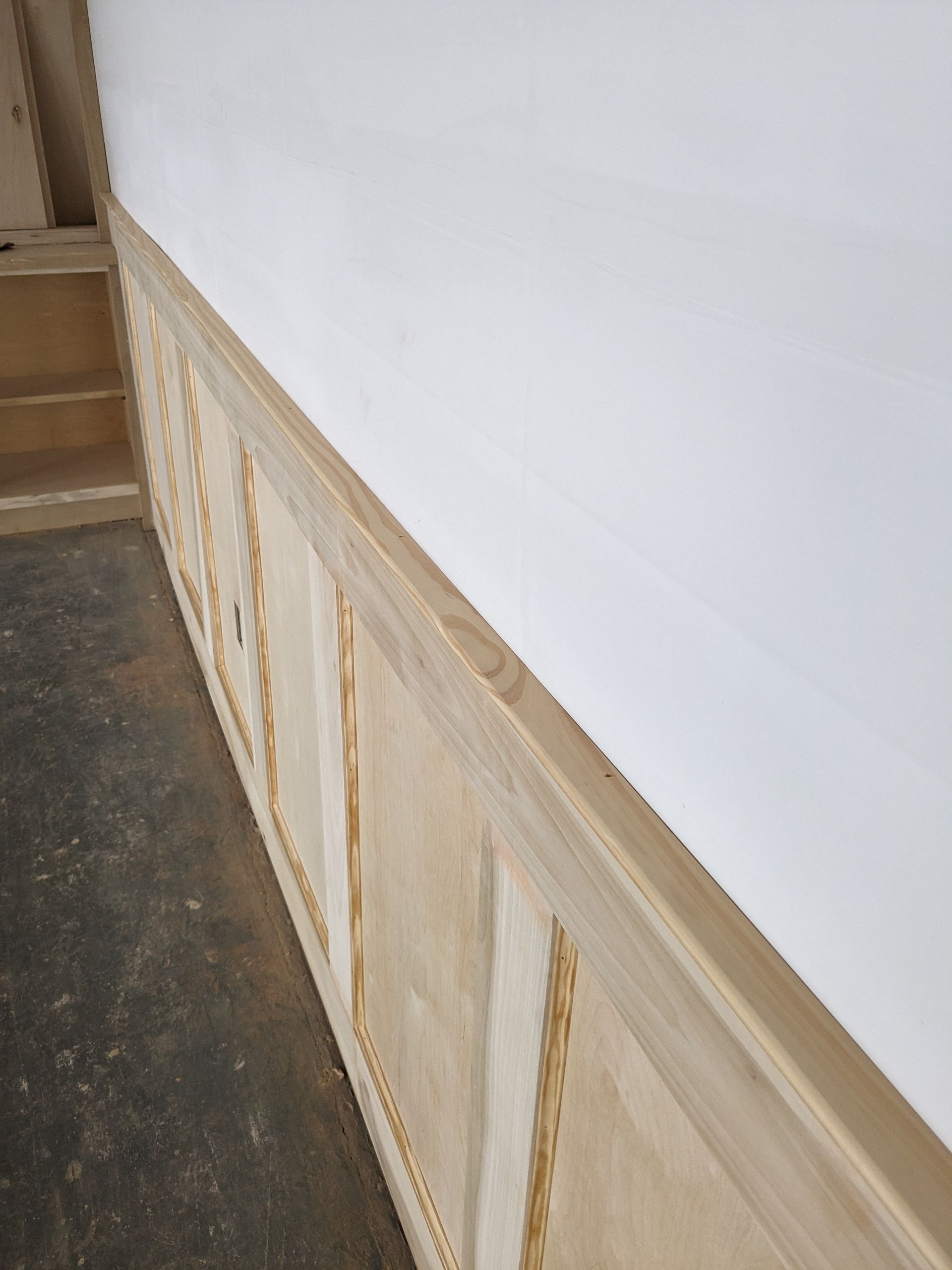 An unfinished interior wall with wooden wainscoting panels installed below a white drywall surface.