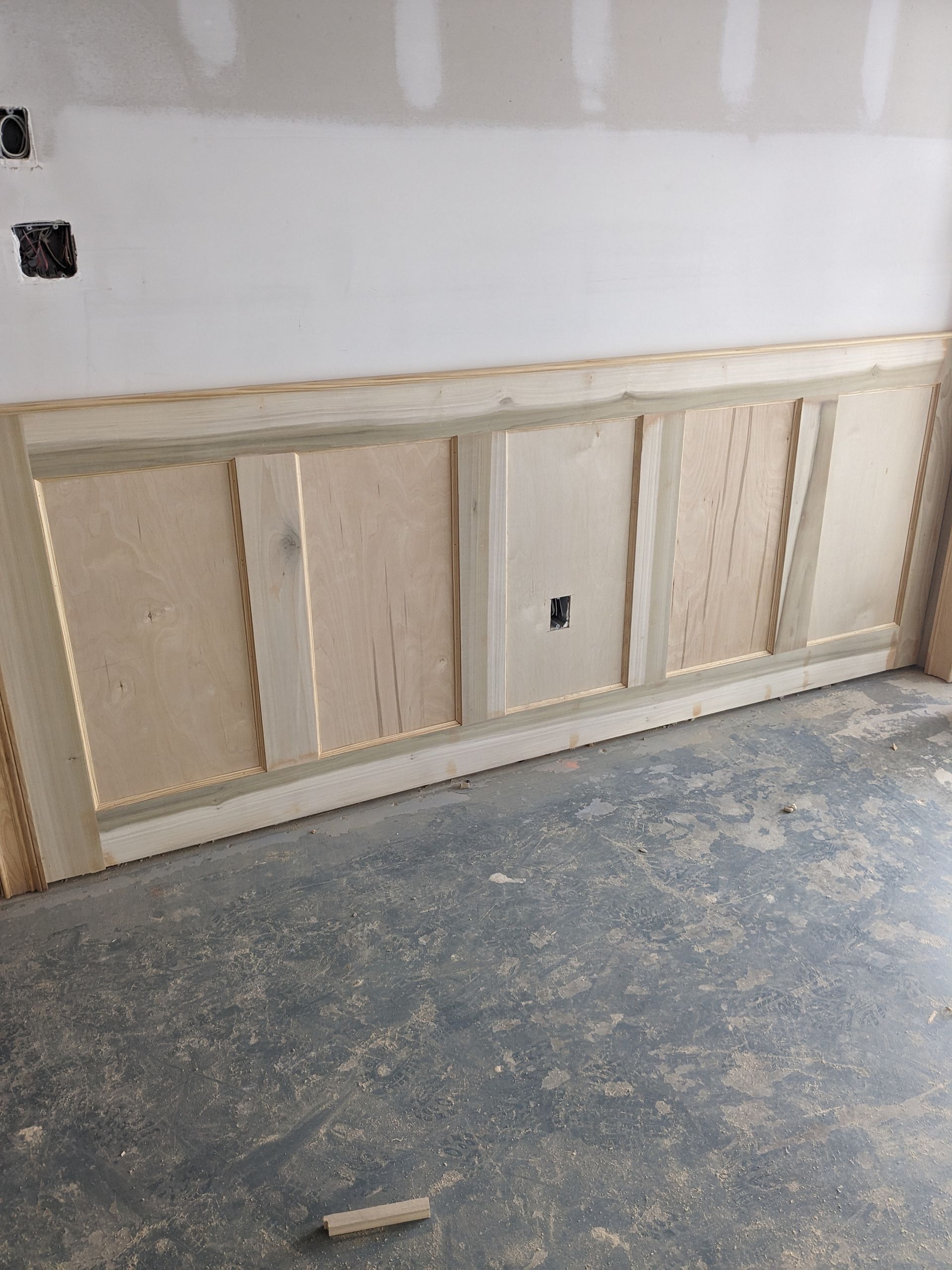 Unfinished wood wainscoting paneling installed on a white wall above a concrete floor in a room under construction.