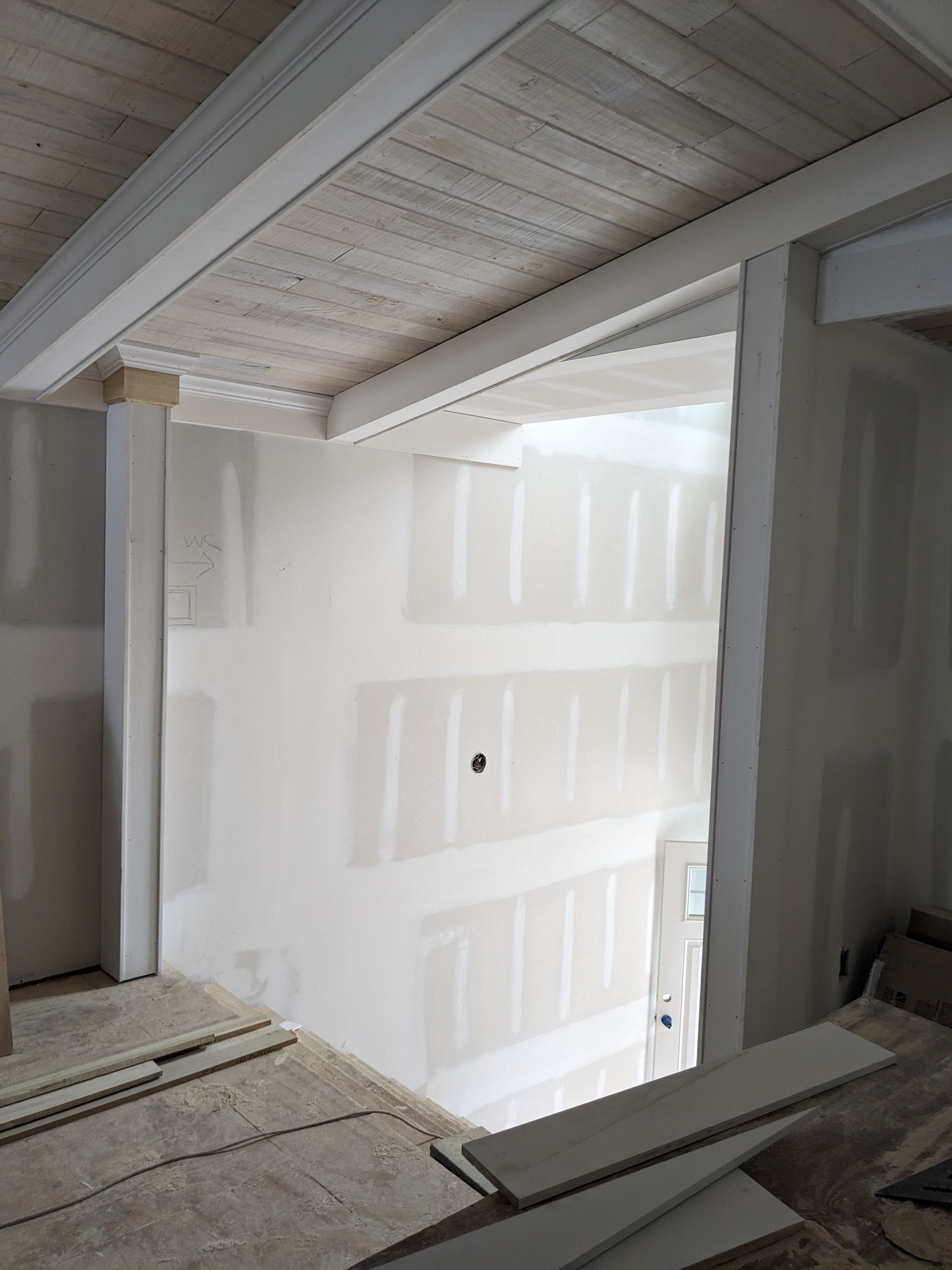 Interior view of a room under construction featuring unfinished drywall, white wooden ceiling beams, and exposed flooring.