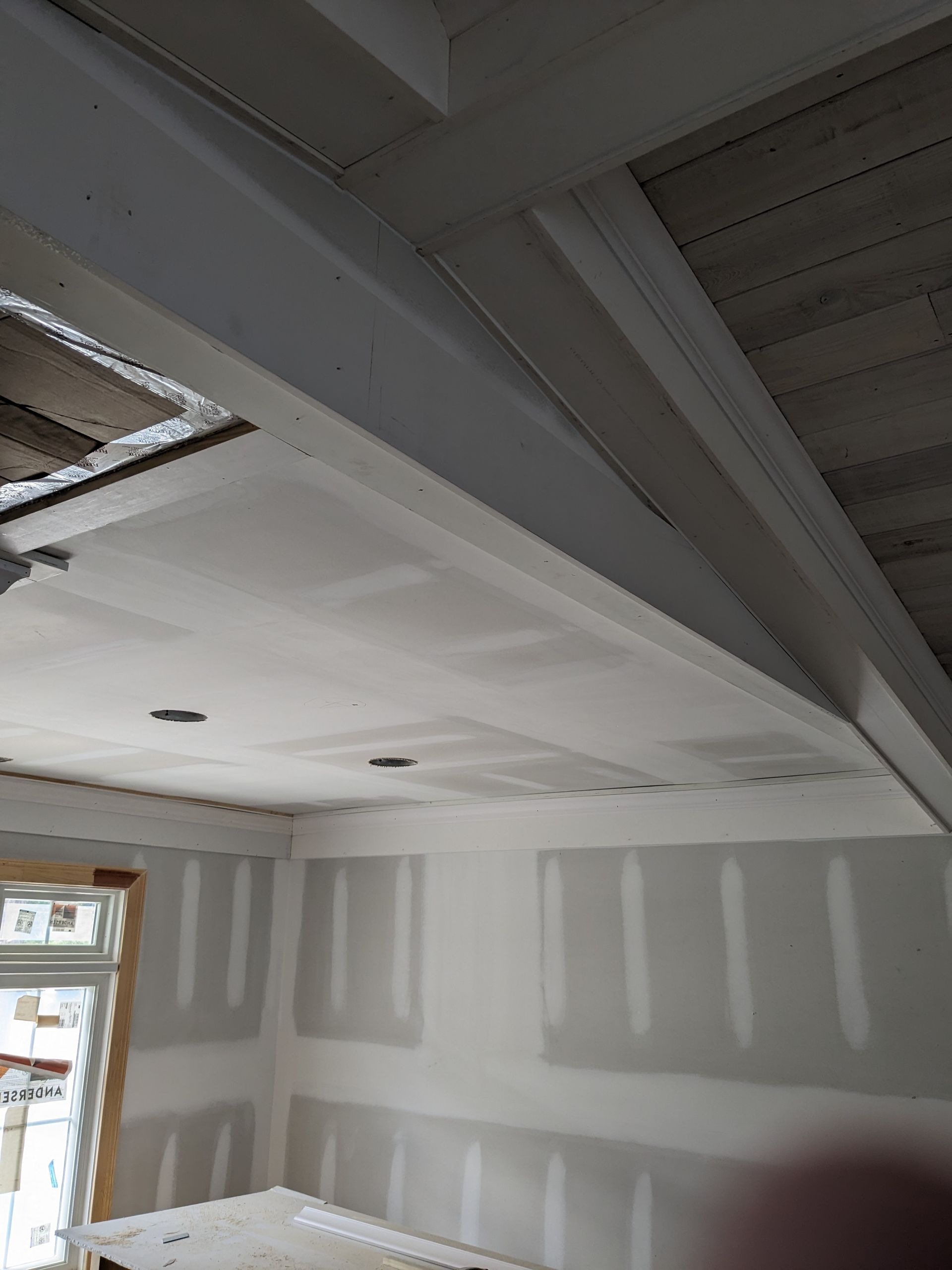 An interior room under construction with white drywall, taped seams, and exposed wooden beams on the slanted ceiling.