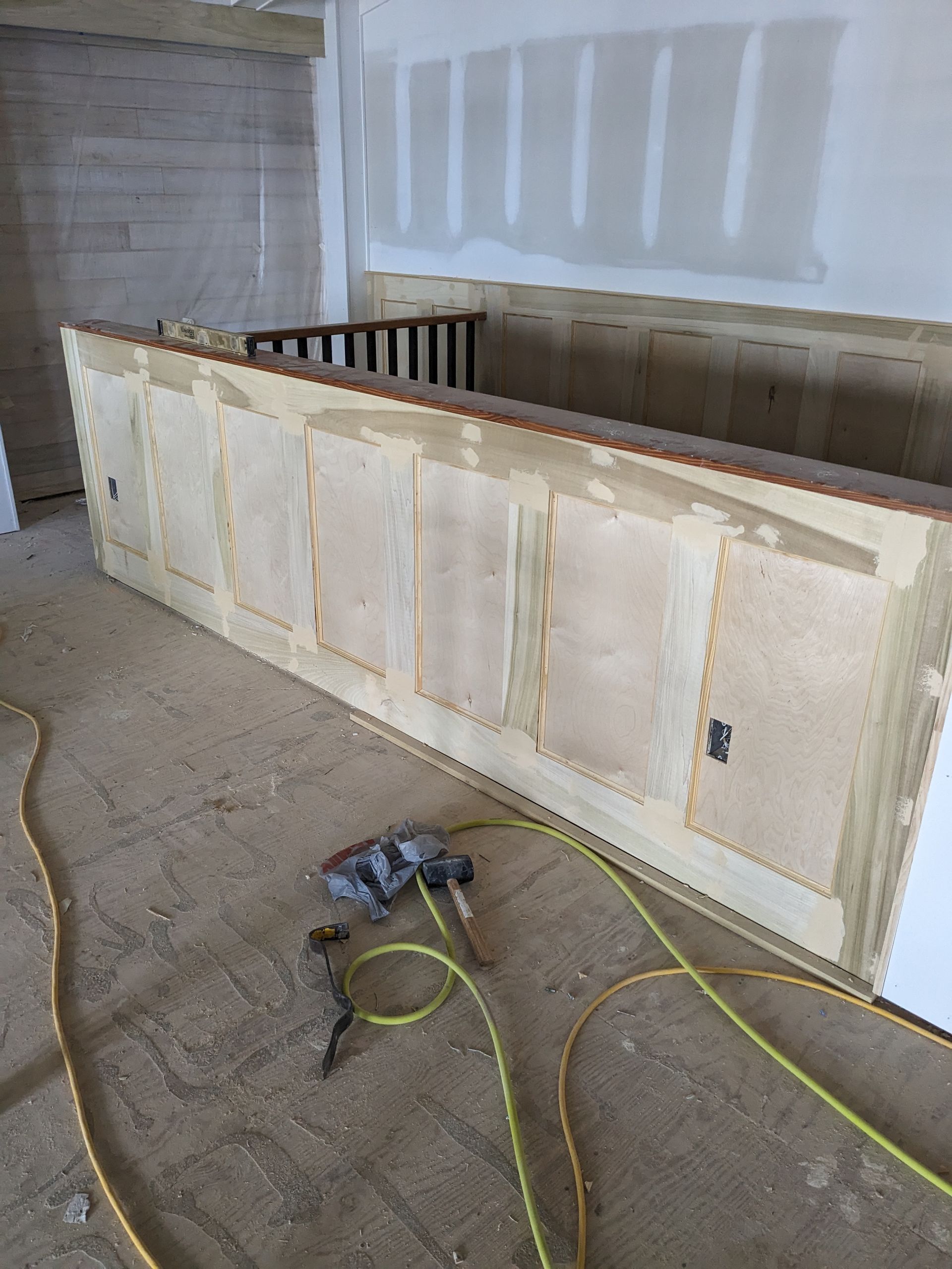 Unfinished wainscoting on a wooden partition wall in a room under construction, with a power drill and cords on the floor.