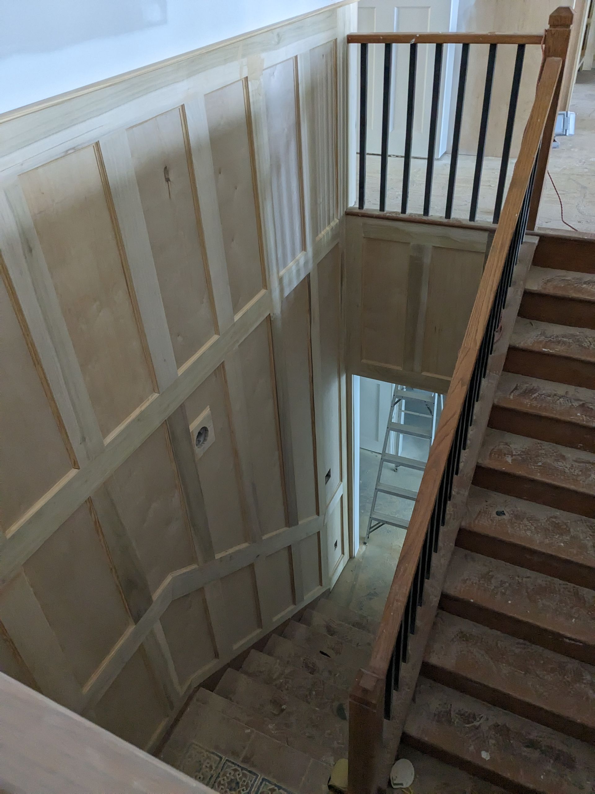 A wooden staircase under construction with wainscoting paneling on the wall and a dark railing.