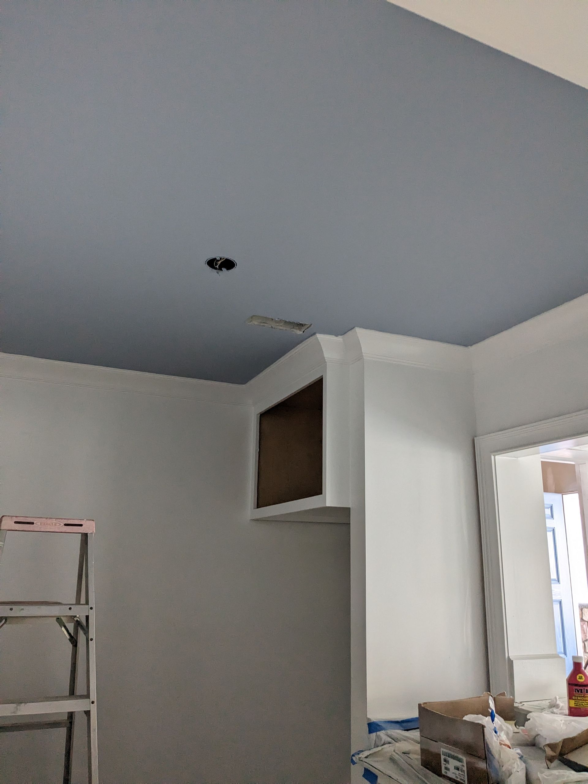 A low-angle view shows a blue ceiling with an electrical box, a white soffit with crown molding, and a ladder to the left.