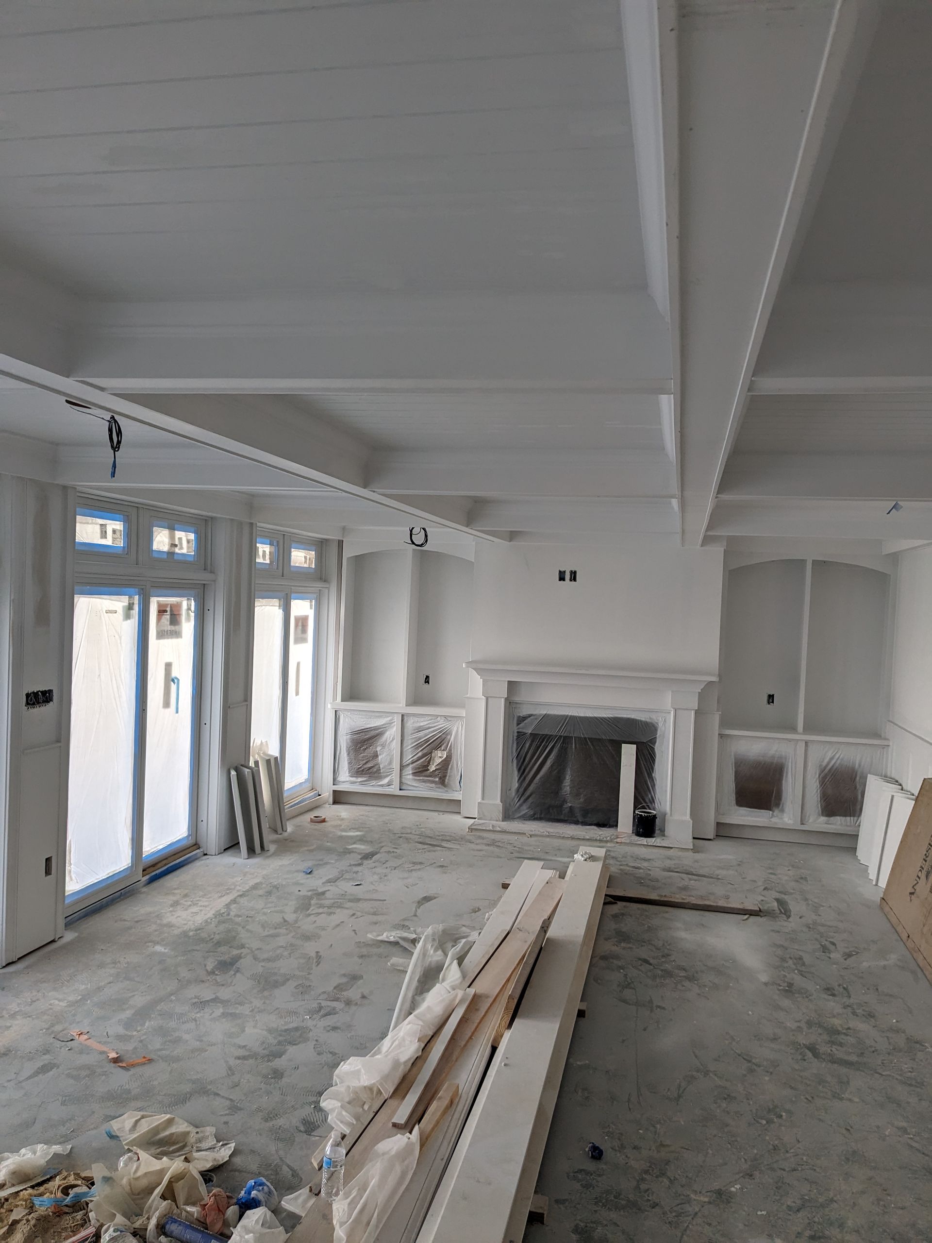 A room under construction with white walls, exposed beams, built-in shelves flanking a fireplace, and glass patio doors.