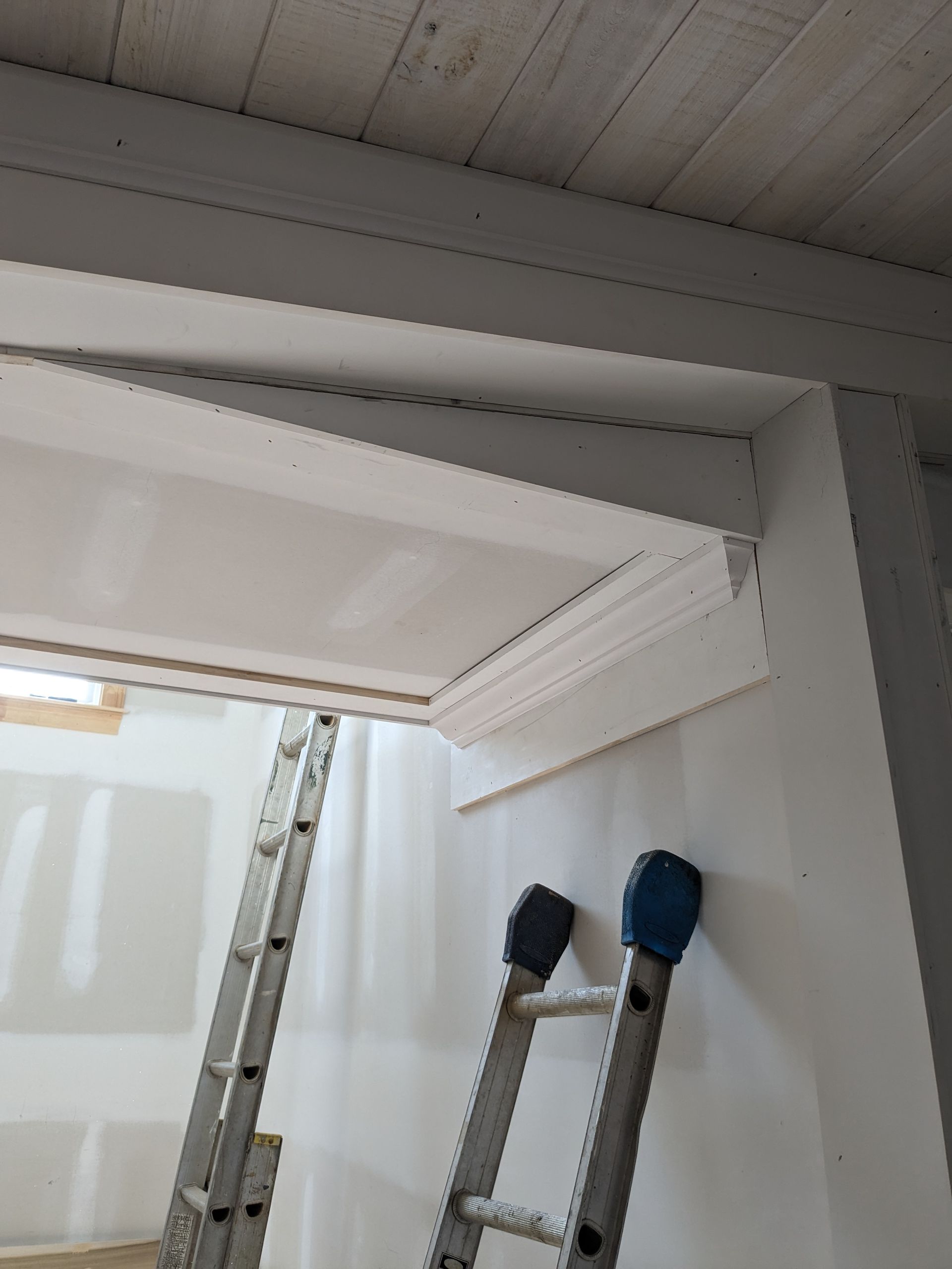 Two metal stepladders stand in a room with white drywall, focused on a newly installed white ceiling beam and crown molding.