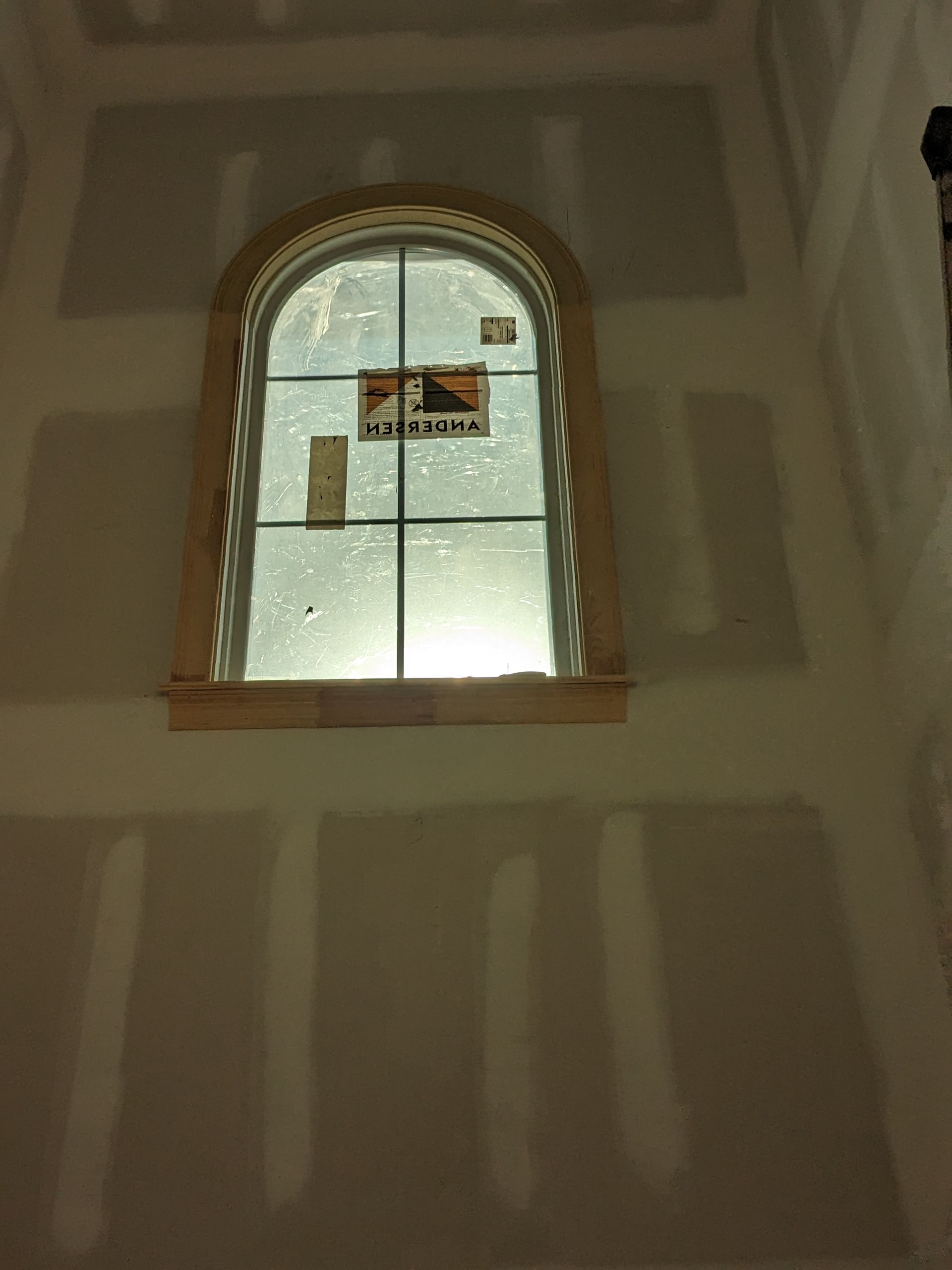 An arched window with wooden trim centered on a drywall-finished wall in an interior construction setting.
