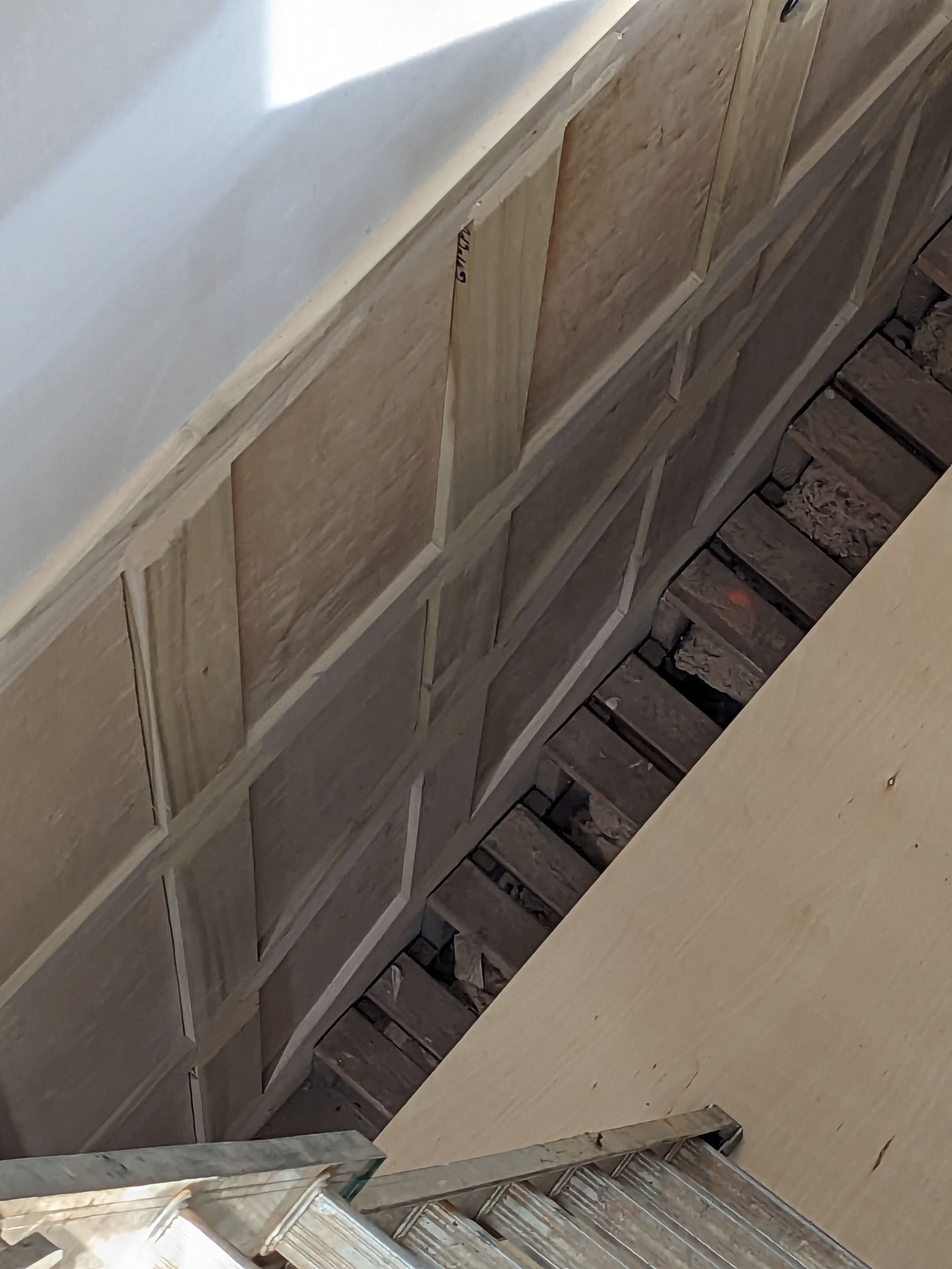 A high-angle view of a wooden paneled wall descending alongside a staircase and an open gap revealing floor joists.