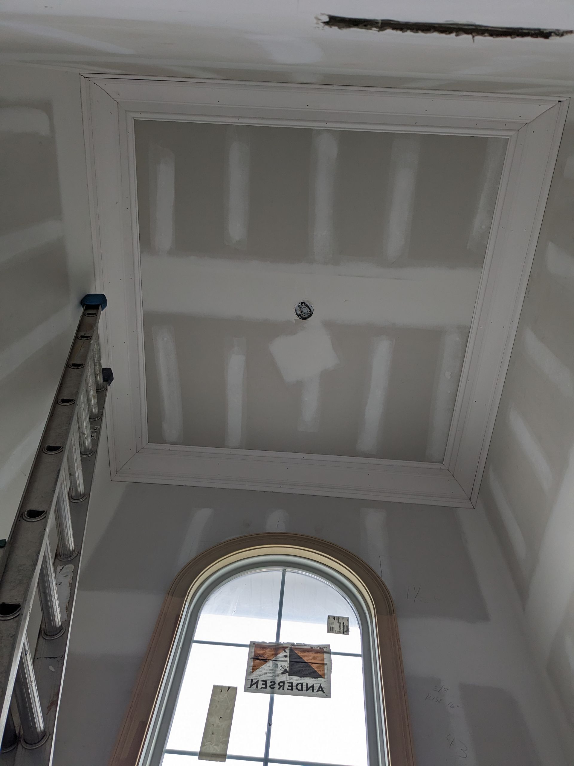 A tall, unfinished interior entryway with gray drywall, a recessed tray ceiling, and an arched window beneath a ladder.