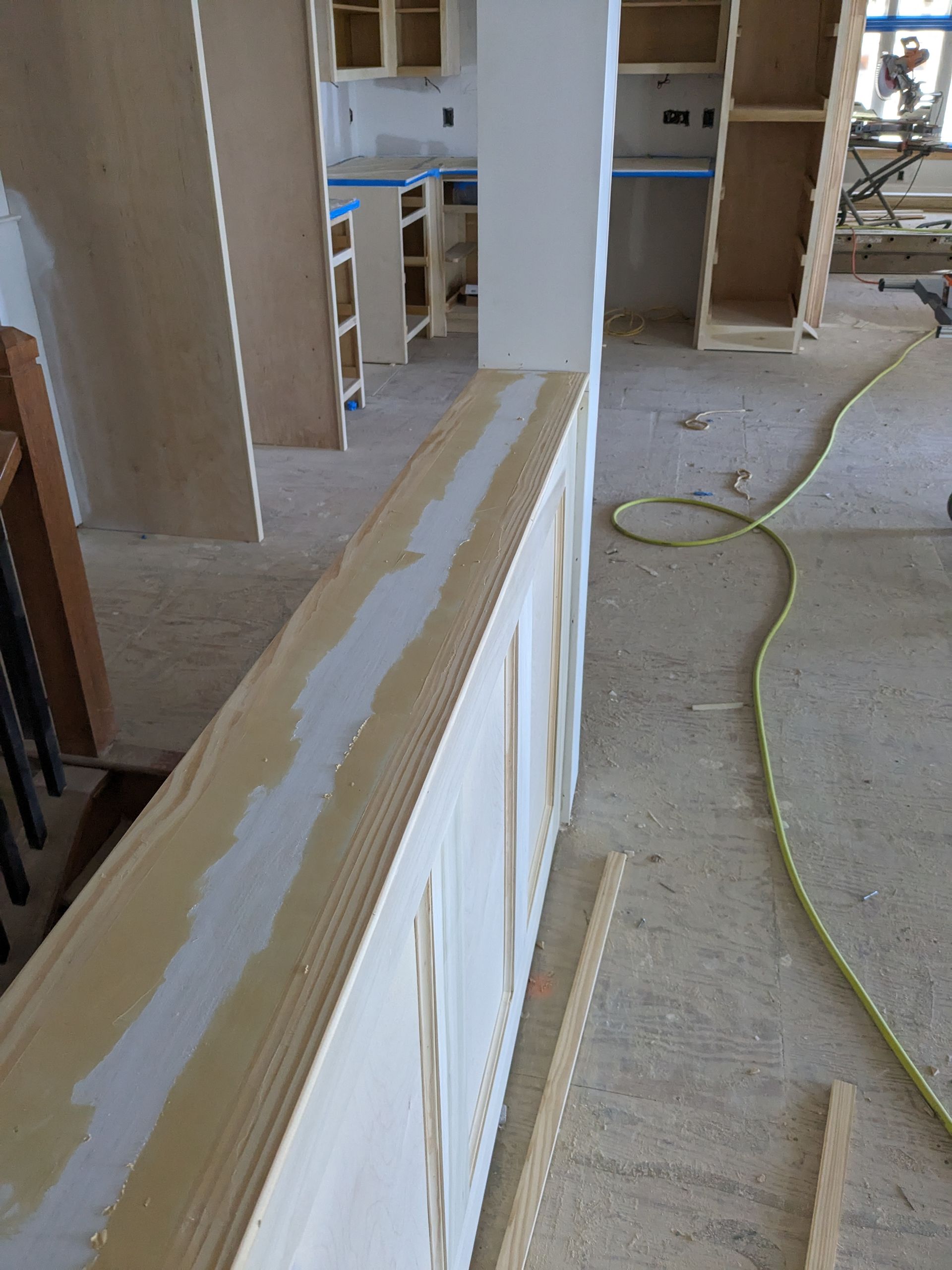 A partially finished half-wall with wainscoting and trim in a room under construction with wood cabinets in the background.