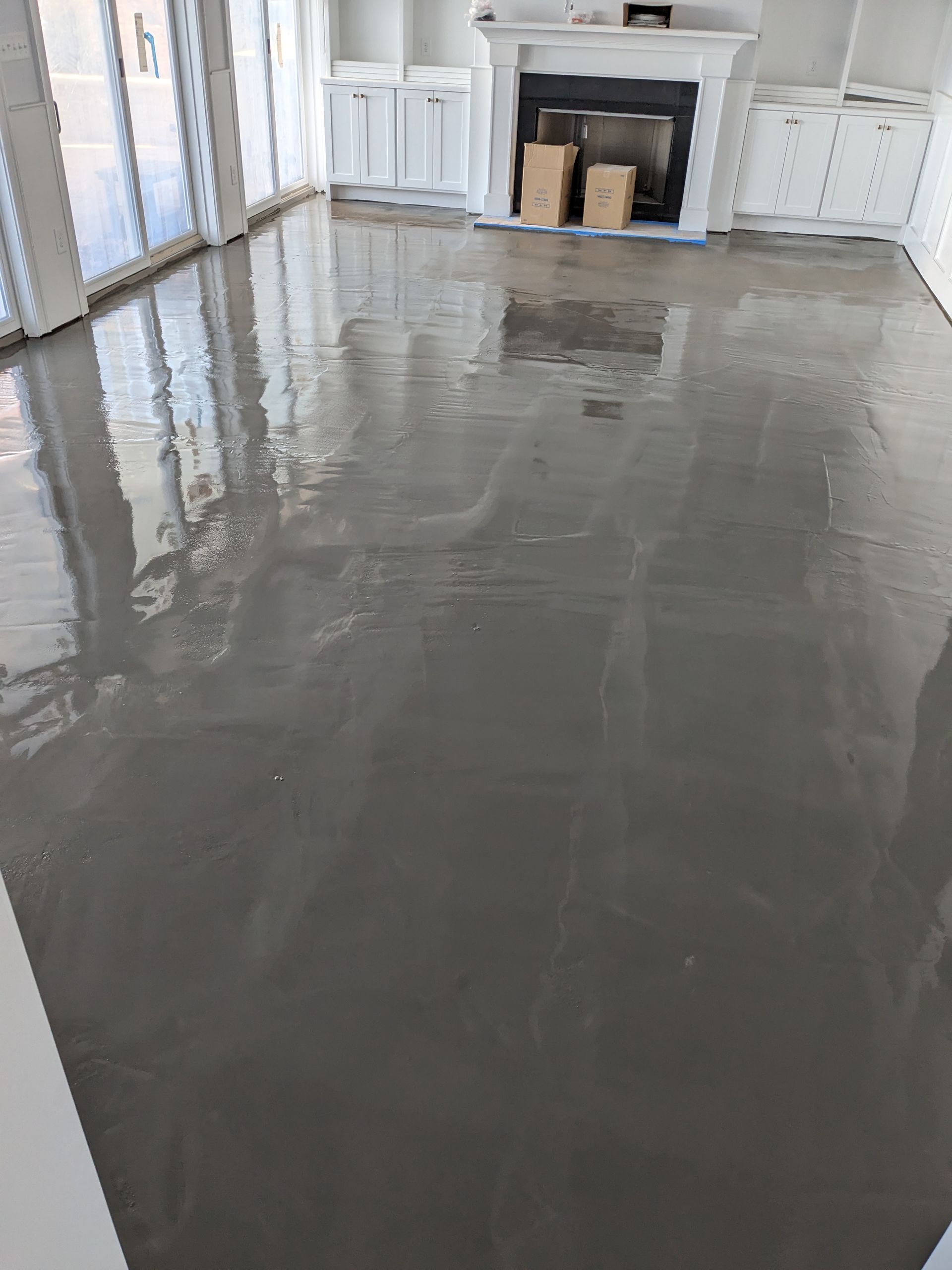 A freshly poured, smooth, gray self-leveling concrete floor in a room with a white fireplace and windowed walls.