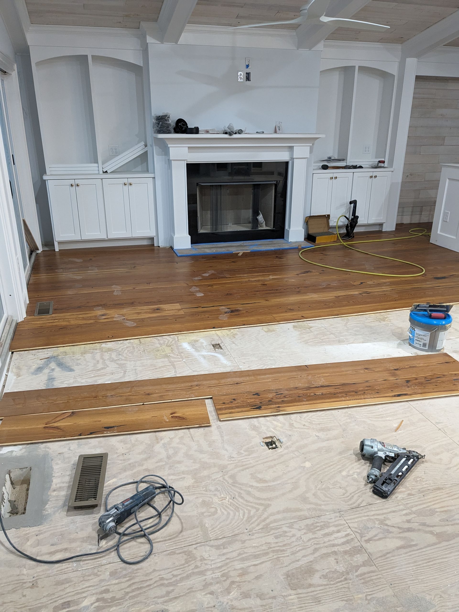 A room under renovation with partially installed hardwood flooring on a plywood subfloor, near a white fireplace.