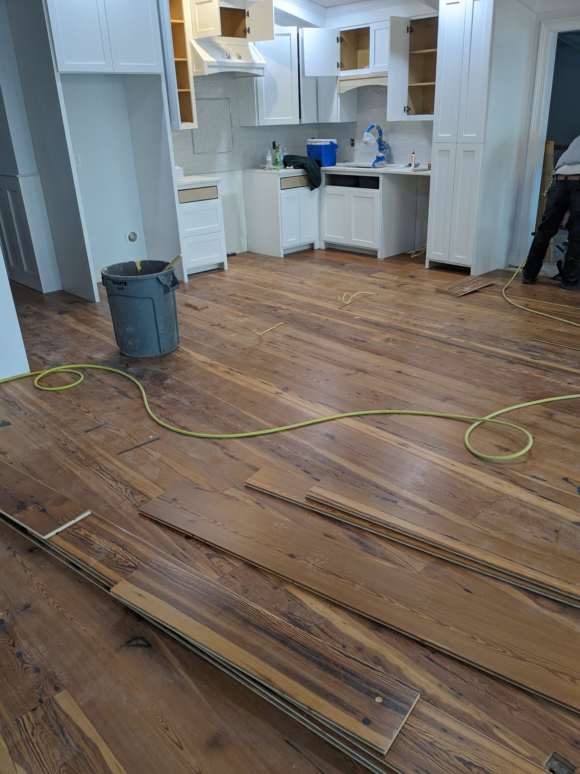 A kitchen undergoing renovation with white cabinets, newly installed wooden floors, and a yellow power cord on the floor.