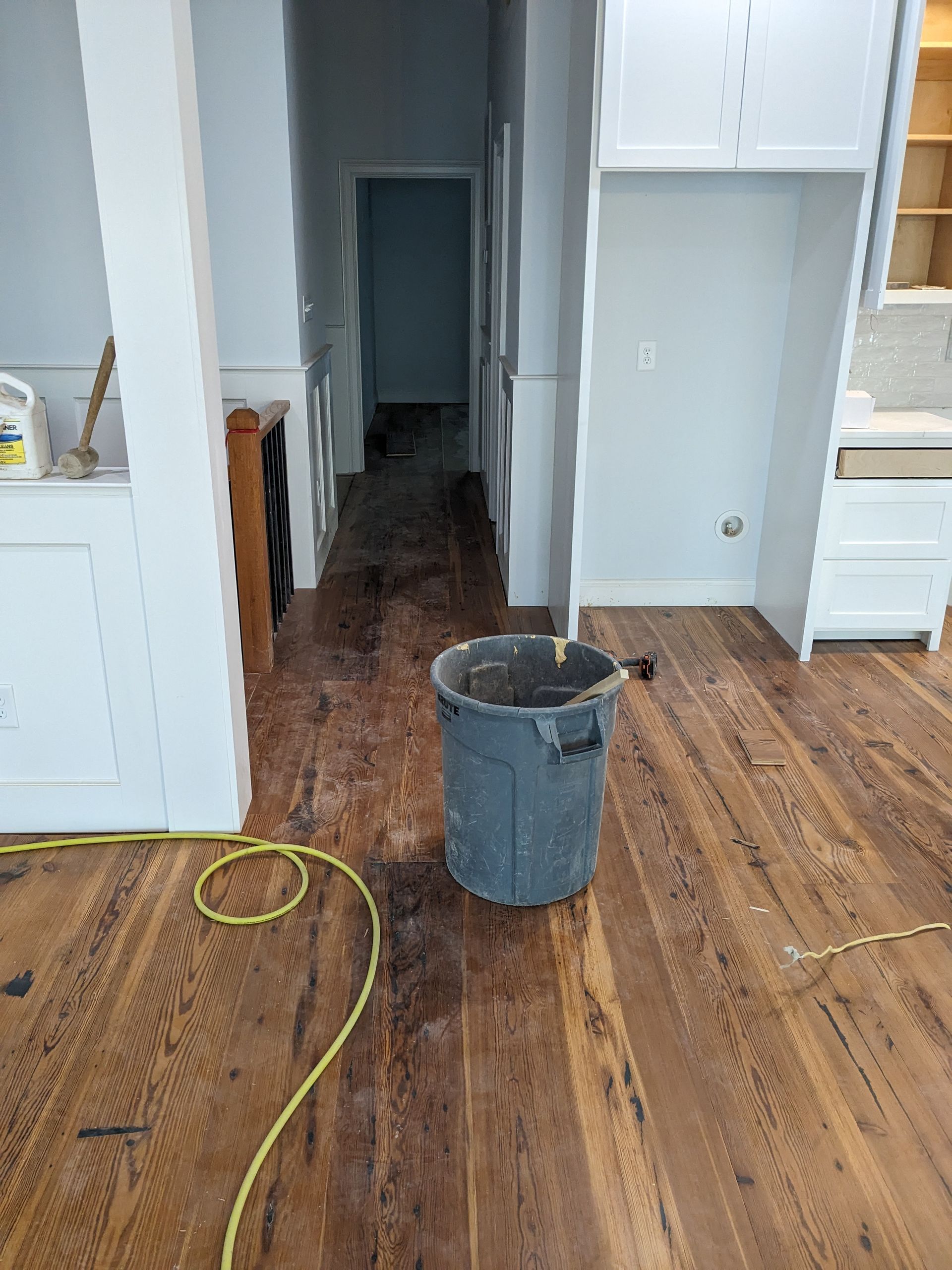 A gray trash can sits in the center of an unfinished room with light wood floors, leading to a dark hallway.